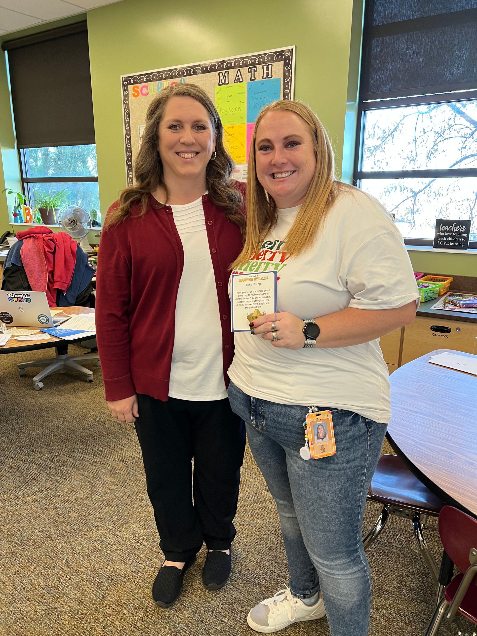 Two women smiling, posing in a classroom. One holds a paper. One wears a maroon cardigan.