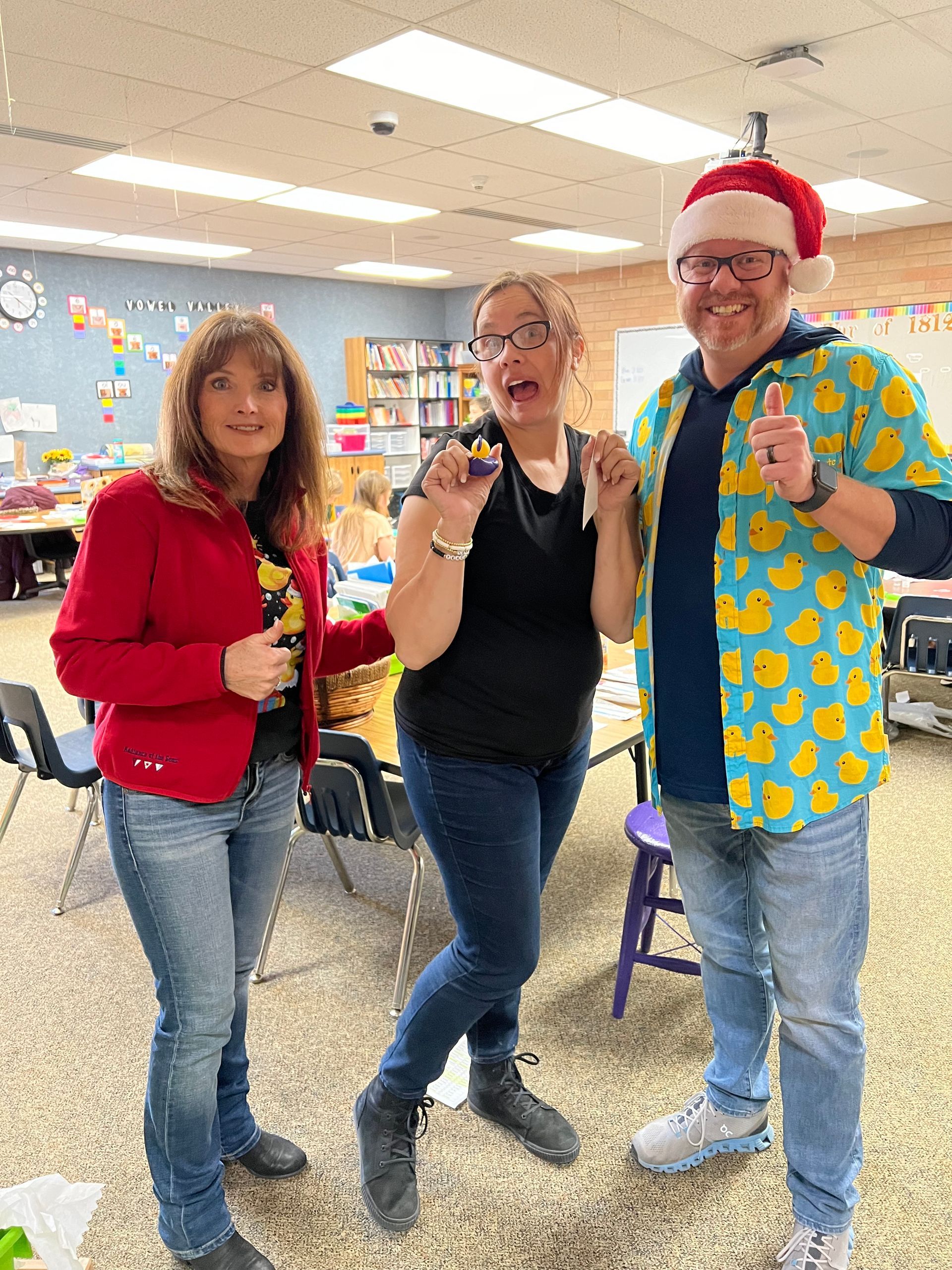 Three people are posing for a picture in a classroom . one of the people is wearing a santa hat.