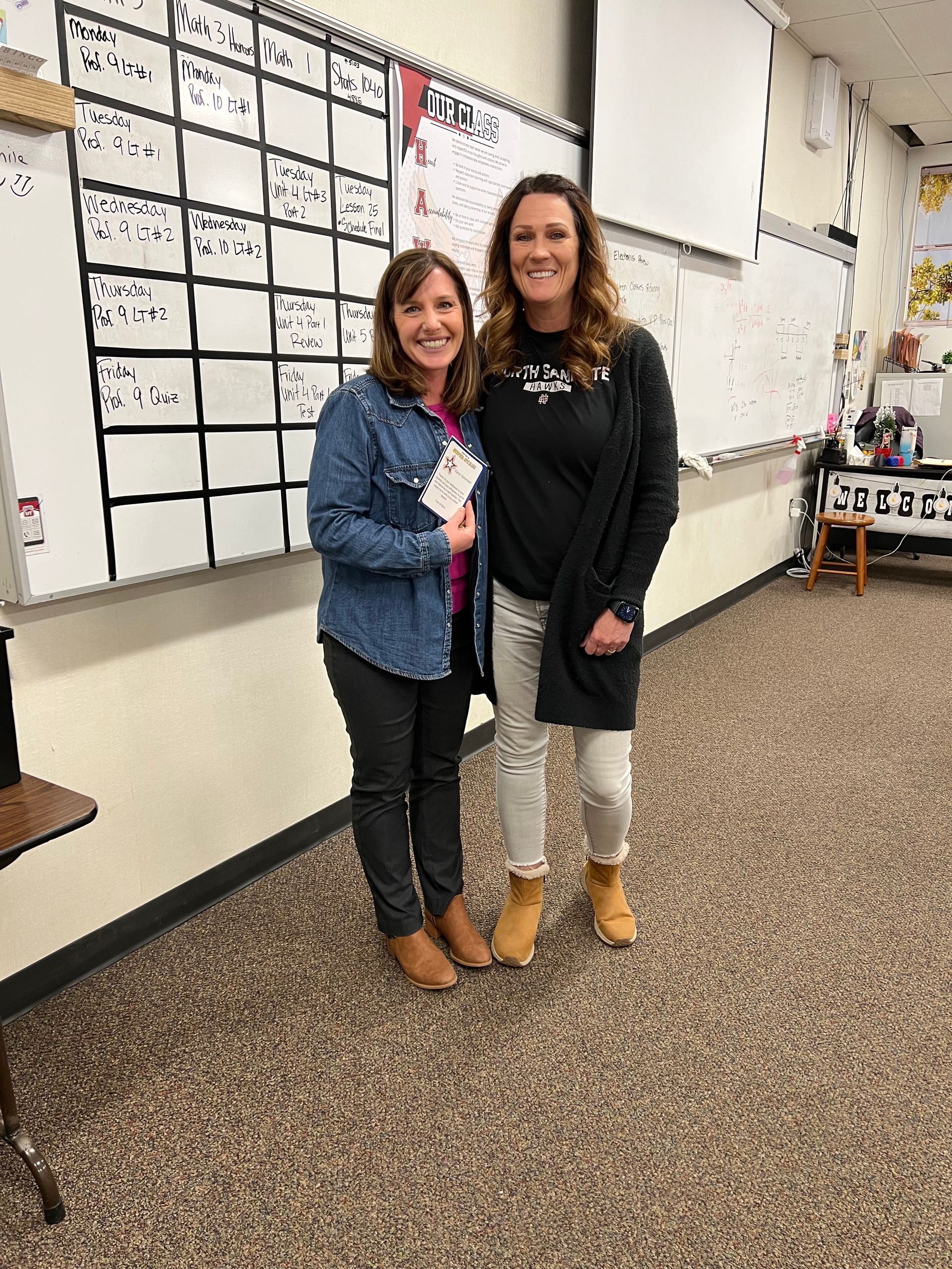 Two women smiling in a classroom with a calendar on the wall. They are wearing casual clothes.