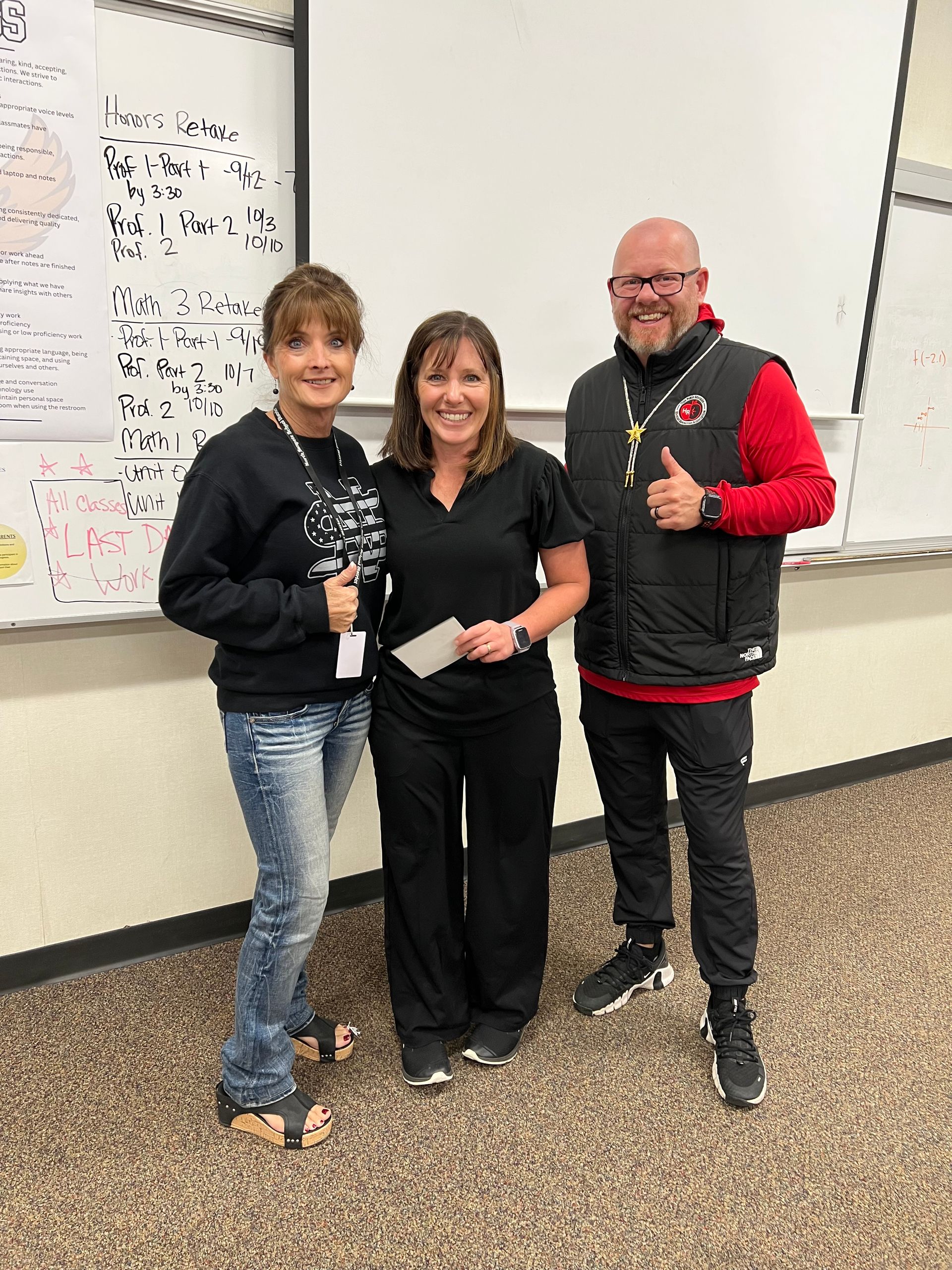 Three people smiling, thumbs up, in front of a whiteboard. One in black scrubs, one in jeans and a sweatshirt, one in a vest.
