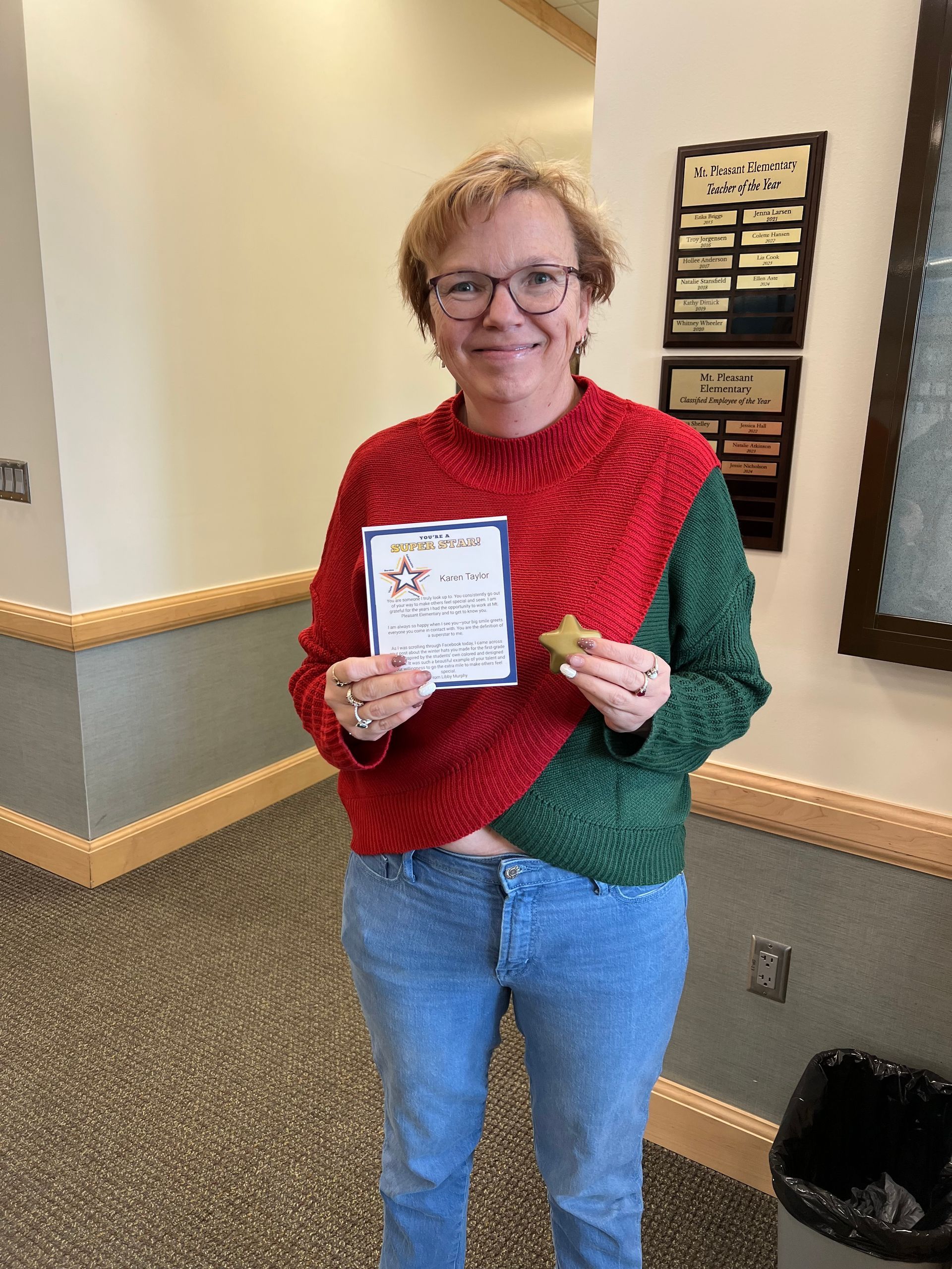 Woman in sweater holding certificate and small item, standing indoors near a wall.