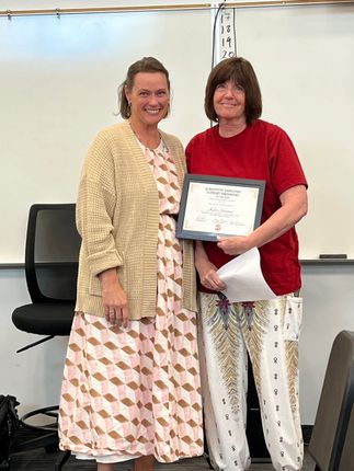 Two women smiling, one holding a framed certificate in a classroom setting