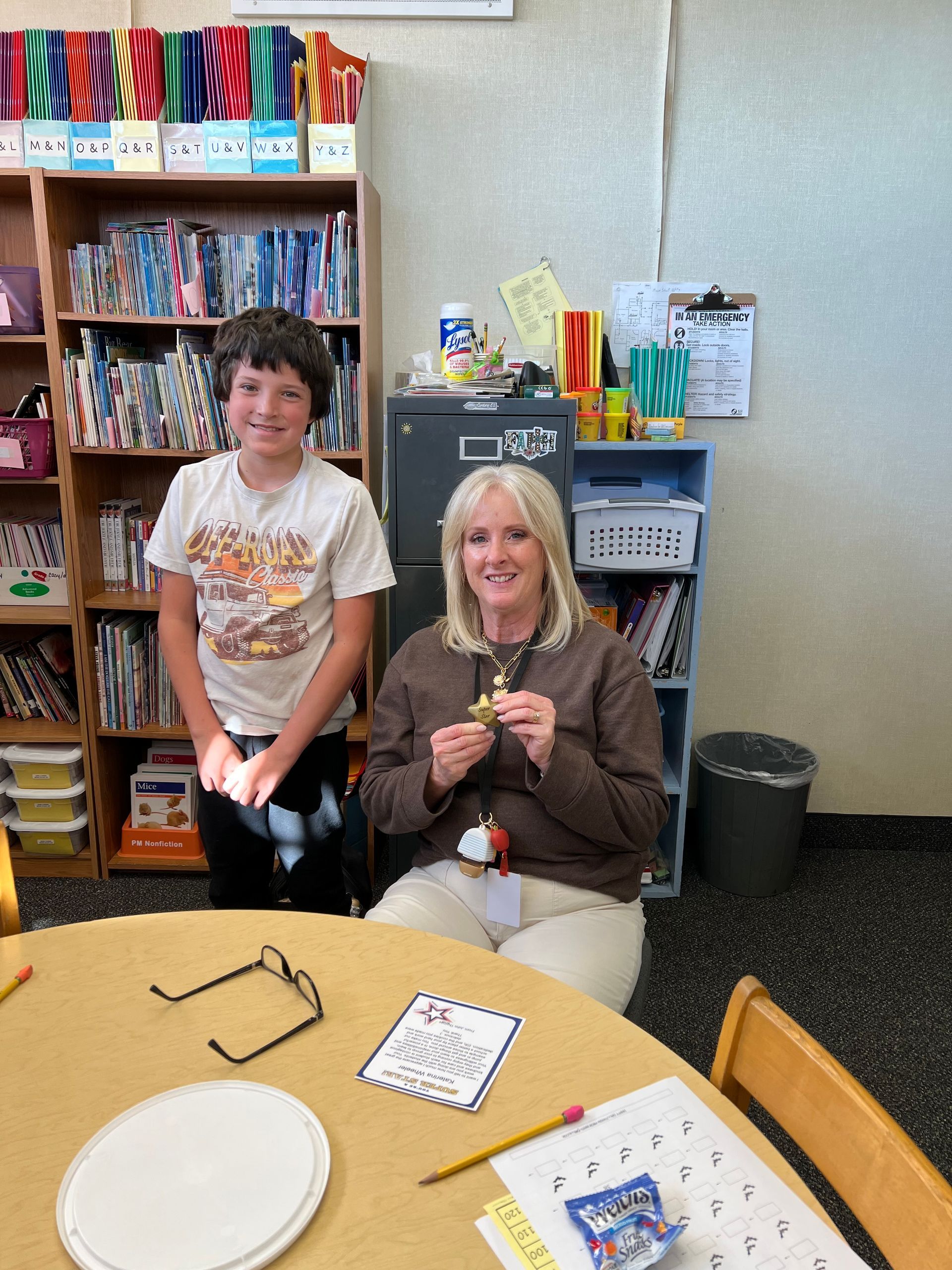 A smiling woman holds an object, sits with a boy at a table. Bookshelves and a bin are in the background.