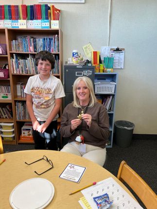 A boy and a woman sit at a table in a classroom. The woman holds a small trophy.