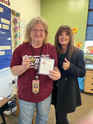 Woman holding award, smiling, with another woman giving thumbs up in a classroom.