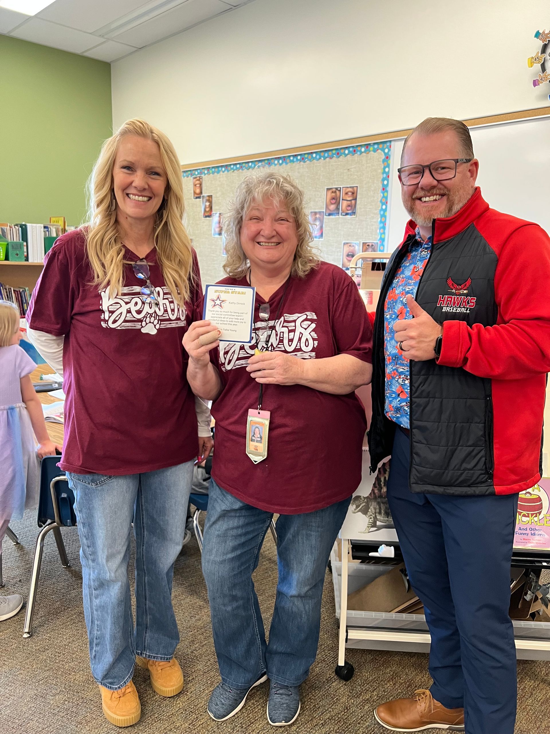 Three people in a classroom, smiling and holding drinks, wearing matching maroon shirts.