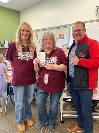 Three people in maroon shirts smiling and holding certificates in a classroom.