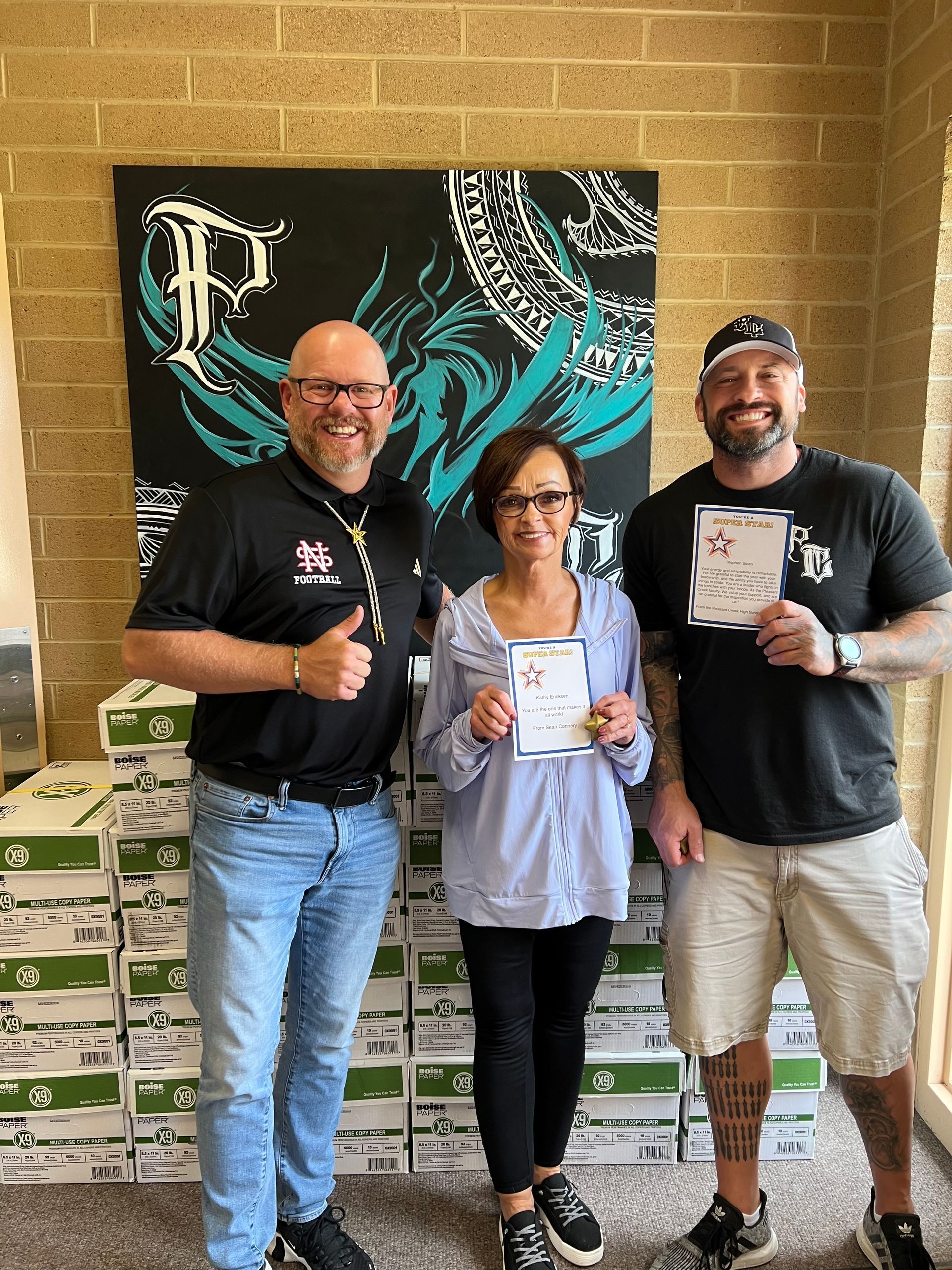 Three people stand with a stack of shoe boxes. They hold a certificate. The man on the left gives a thumbs up.