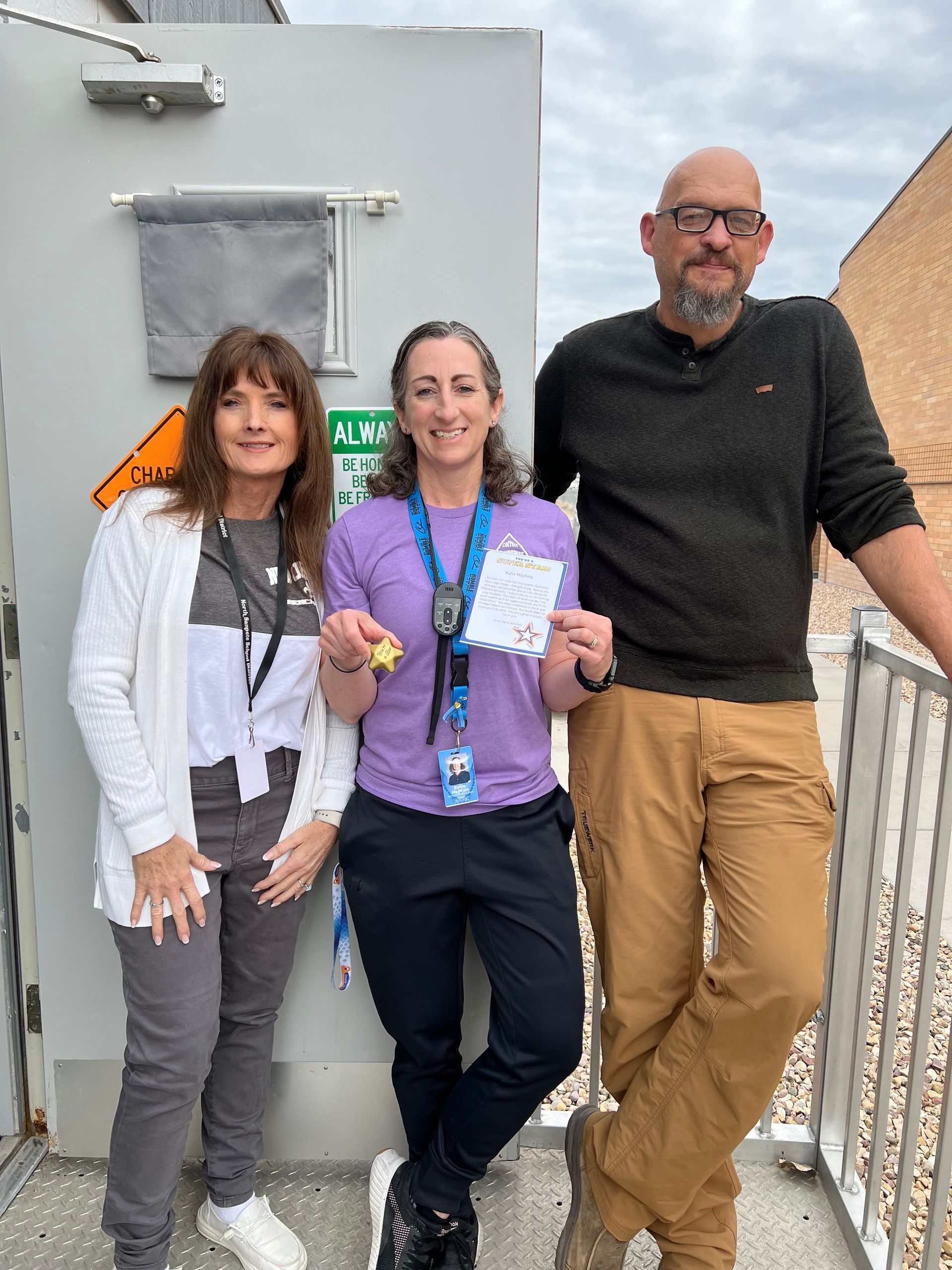Three people stand outdoors near a wall, smiling and posing for a photo. The center person holds a small card and a charm.