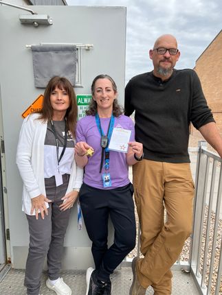 Three people stand outdoors near a wall, smiling and posing for a photo. The center person holds a small card and a charm.