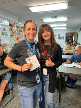 Two women in a classroom; one holding an award and smiling, the other with a thumbs-up. Students in background.