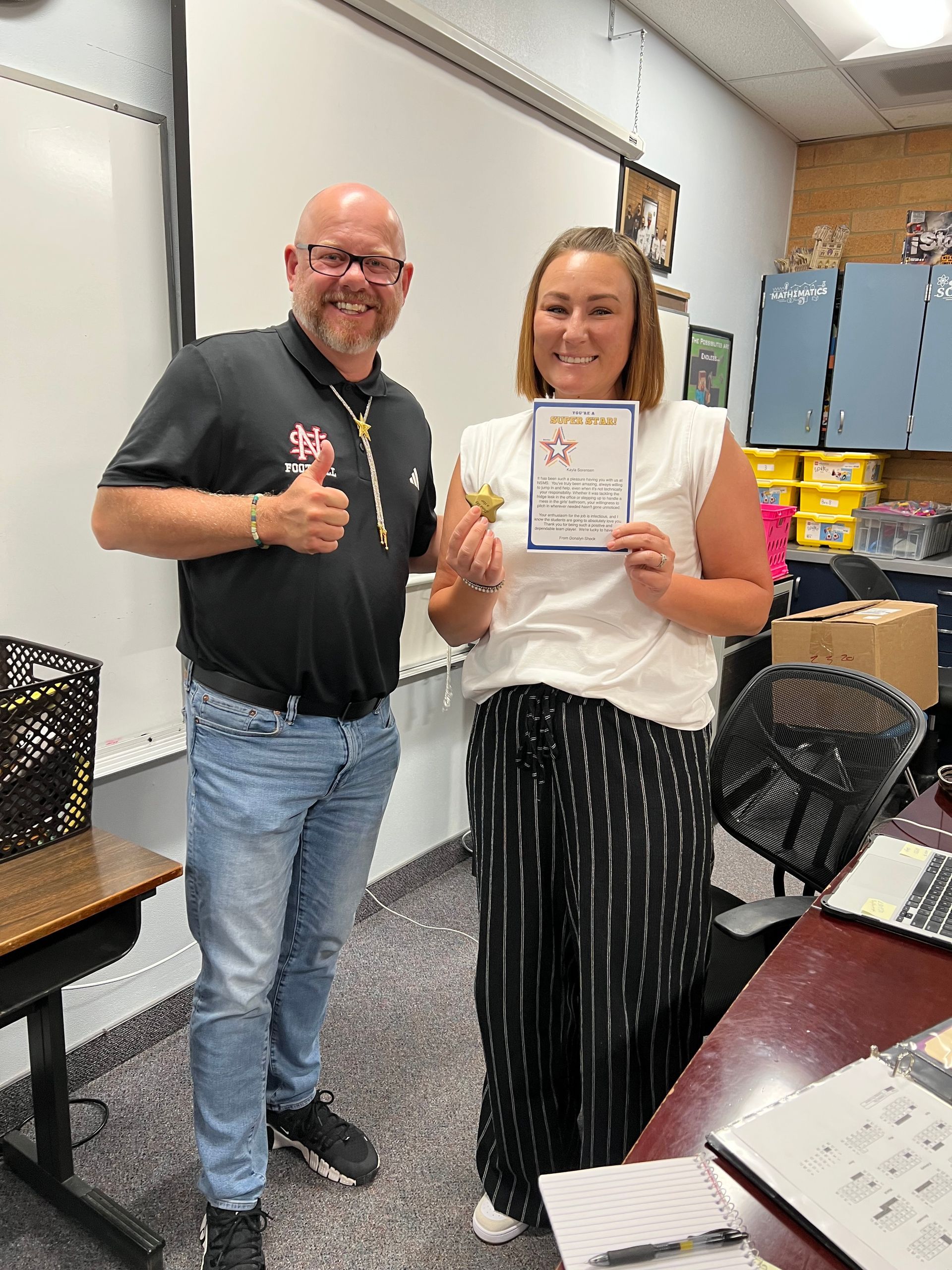 Man giving thumbs up, woman holding certificate, in classroom.