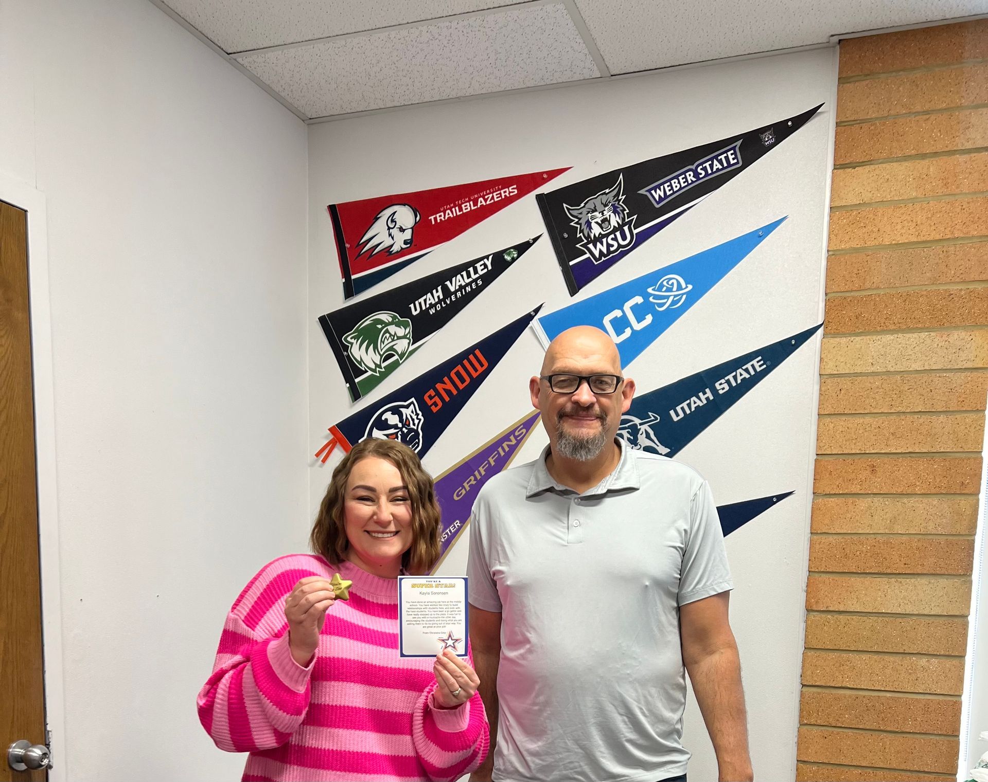 Woman in pink sweater and man in gray shirt pose by pennant-decorated wall.
