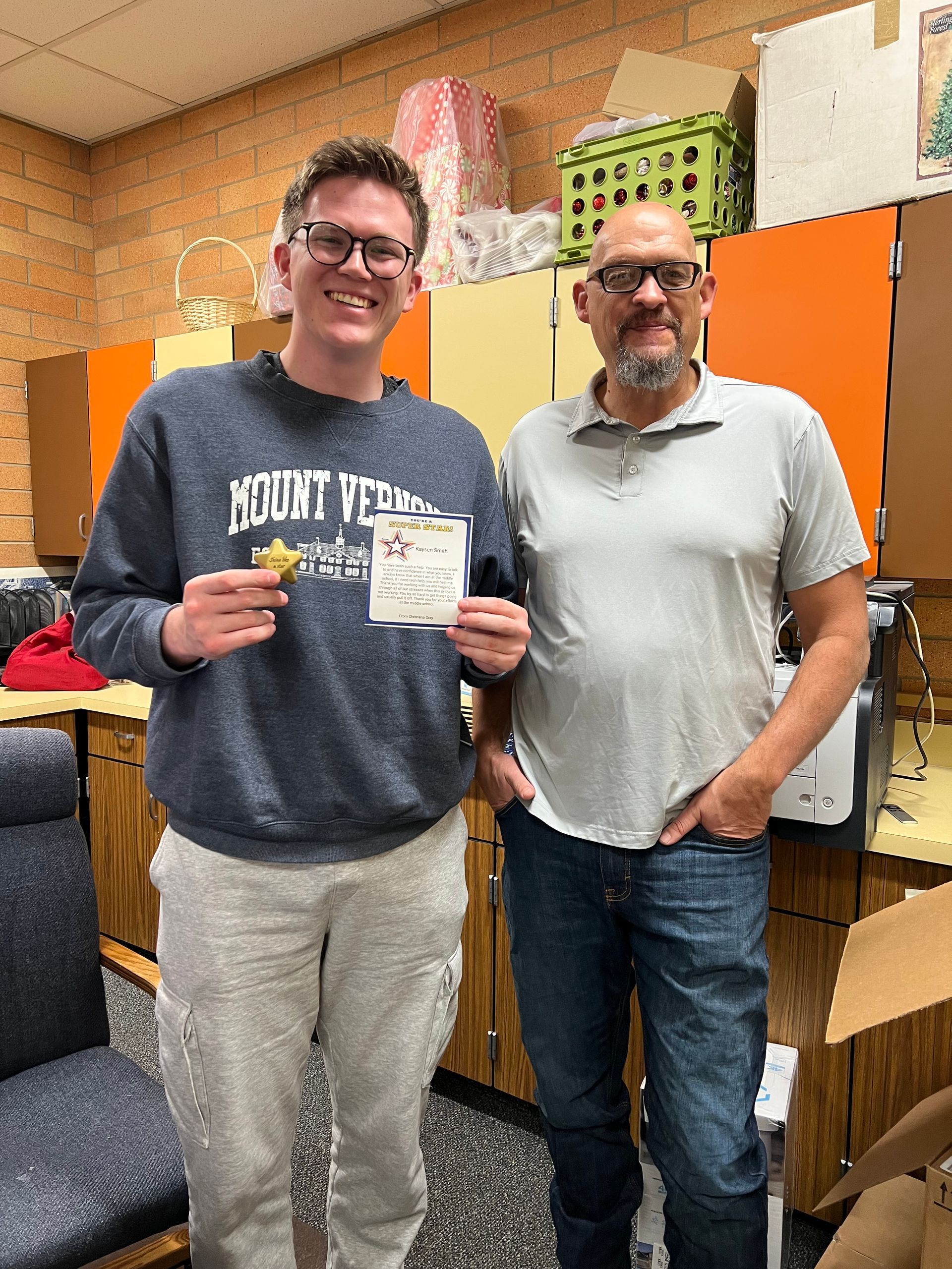 Two men in a classroom holding a star and paper. One in a blue sweatshirt, the other in gray.