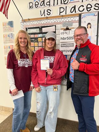 Three people in red shirts smiling and holding a certificate in front of a classroom bulletin board.