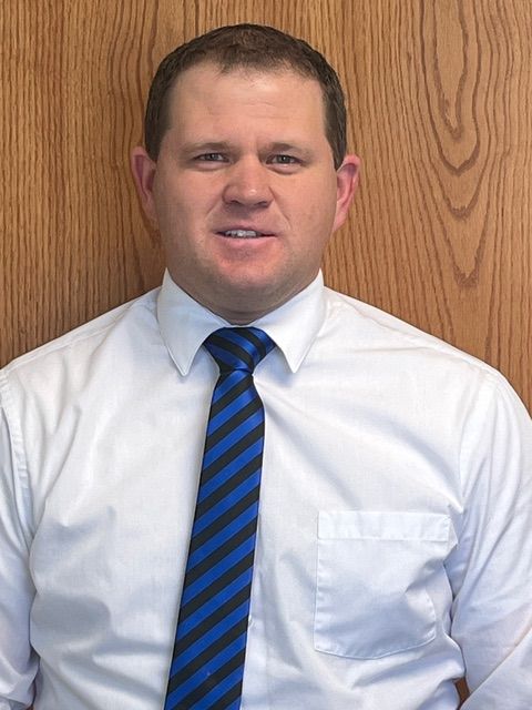 Man in white shirt and blue striped tie smiles, wood panel background.