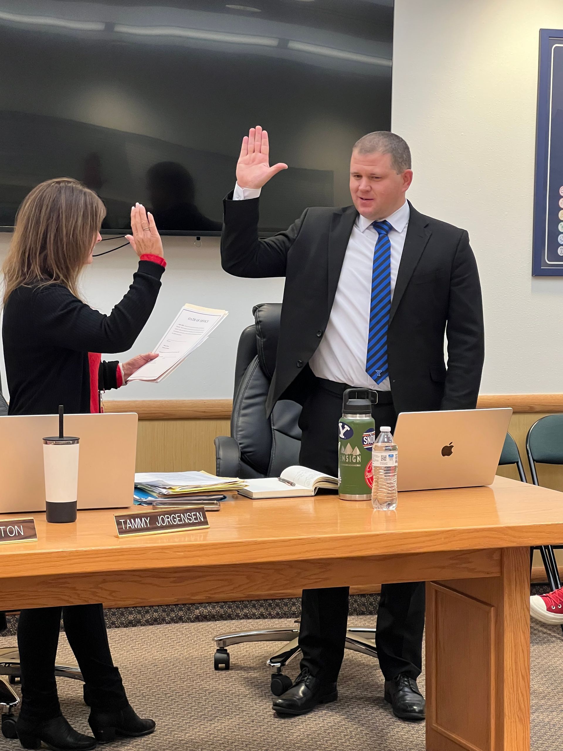 Man in suit raises hand, being sworn in, another person giving him a high-five at a desk.