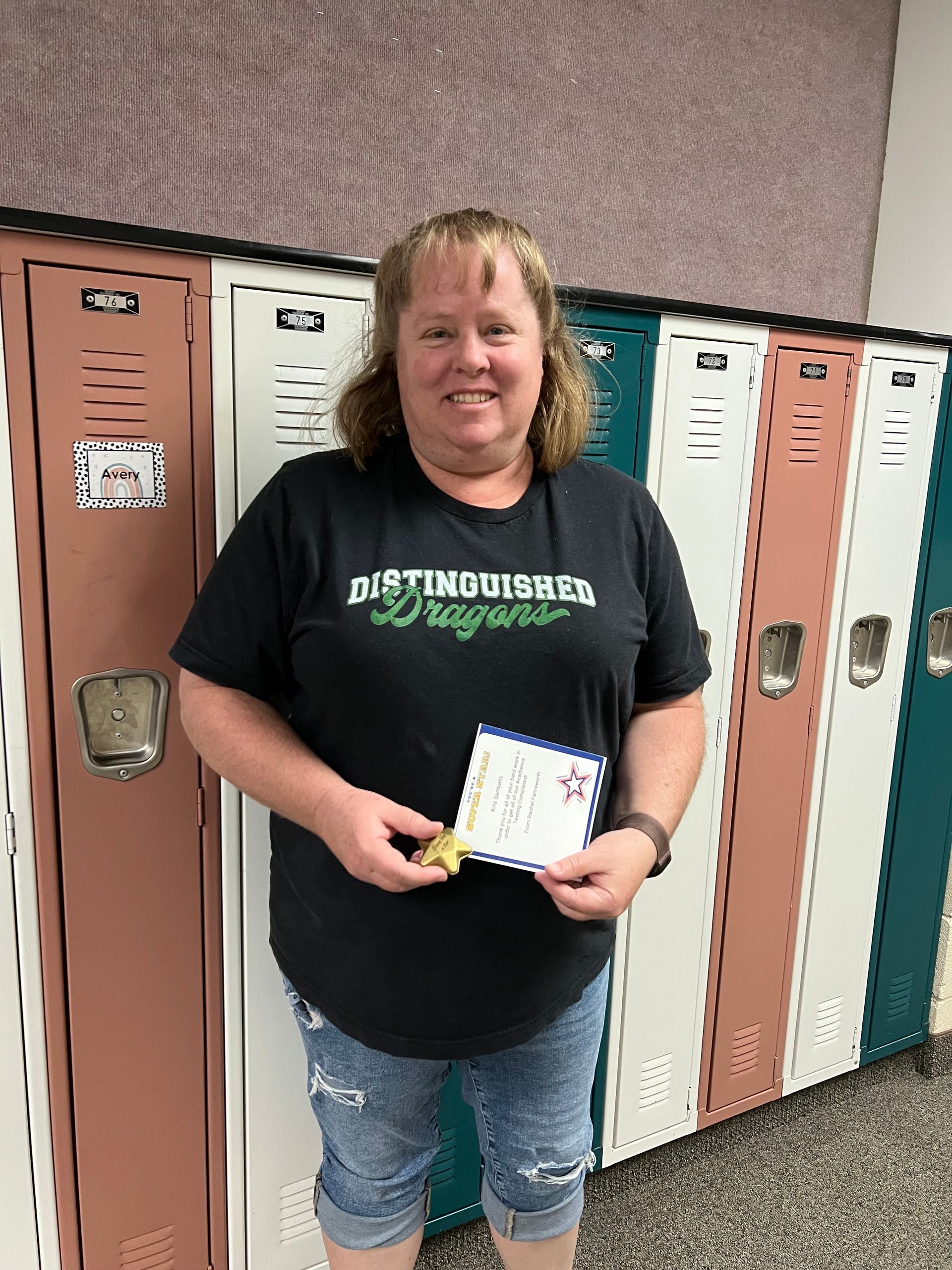 Woman stands by lockers, smiling, holding a certificate and star.