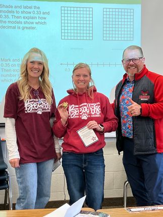 Three people standing indoors in front of a whiteboard, smiling and holding papers.