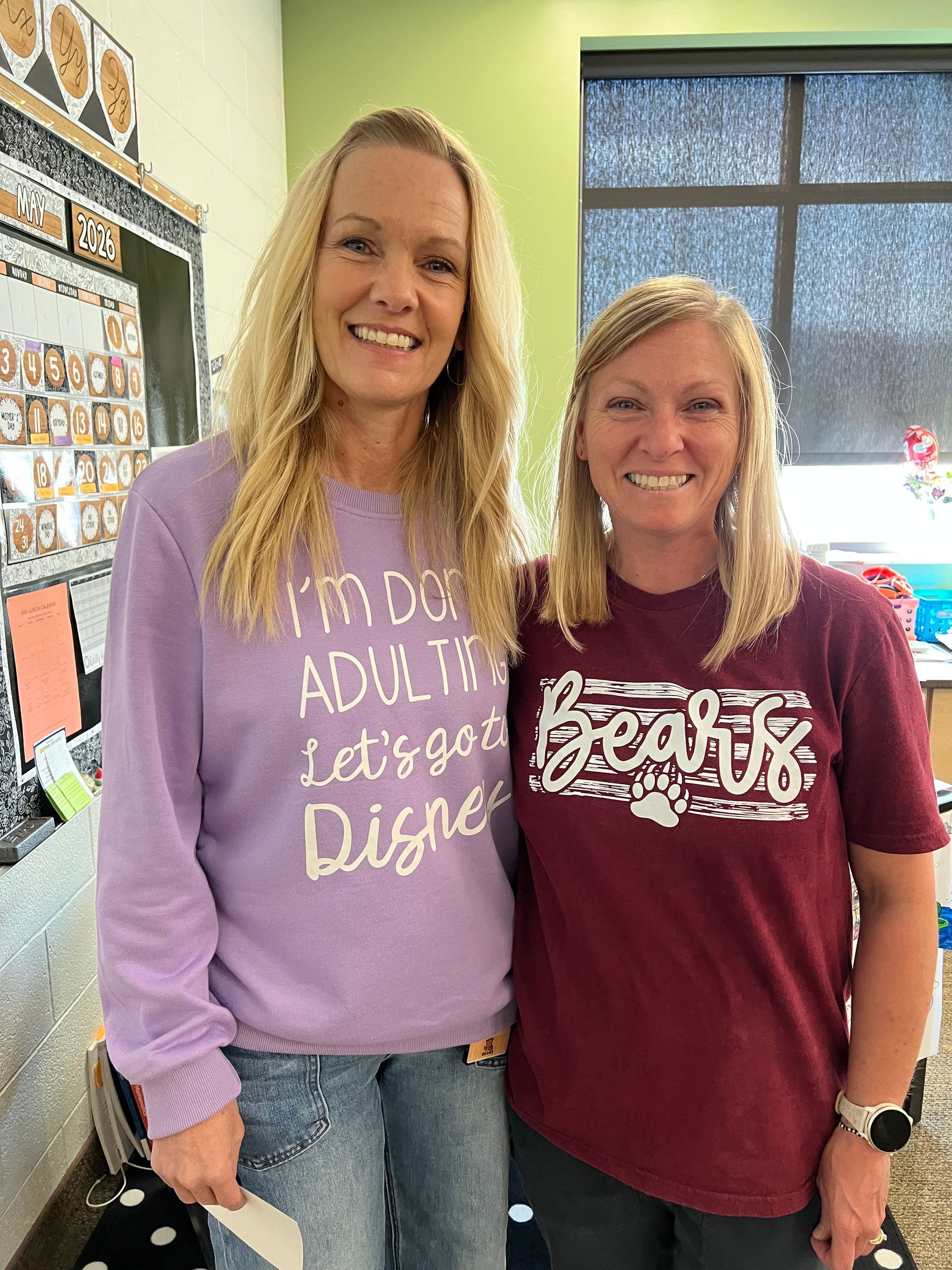 Two smiling people standing together indoors, wearing purple and maroon sweatshirts, in a classroom.