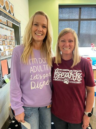 Two women smiling indoors, standing side by side in casual sweaters.