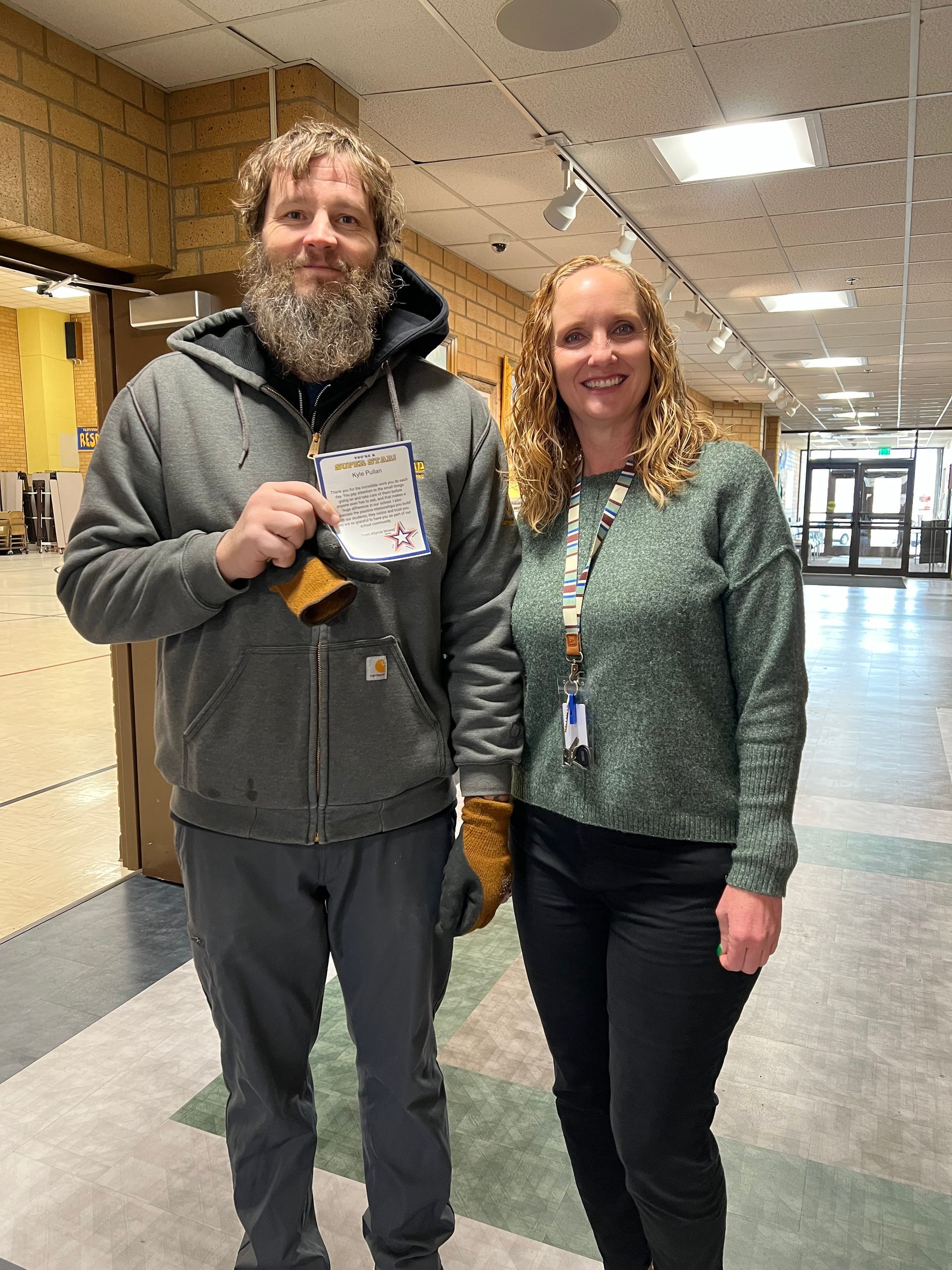 Man with beard and woman in a hallway. Man holds paper and wears a coat. Woman wears sweater and pants.