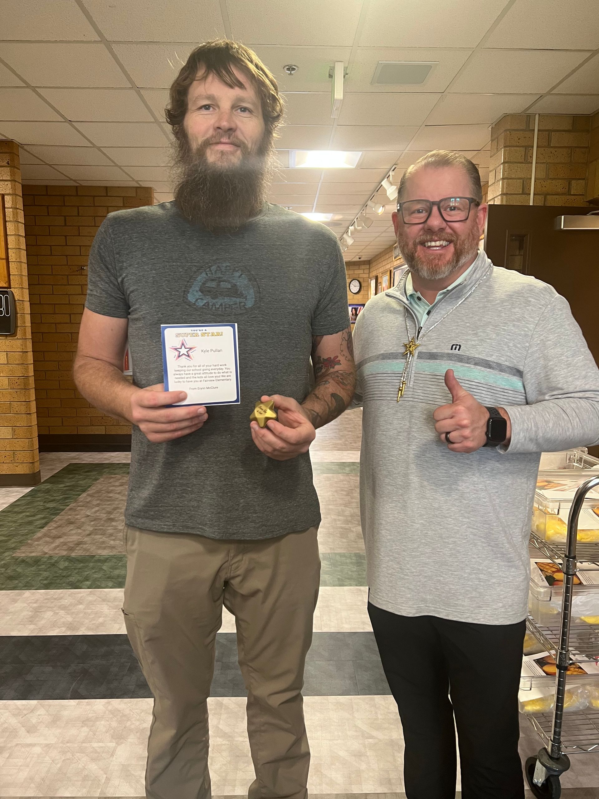 Two men standing indoors, one holding an award and a gold coin, the other giving a thumbs up.