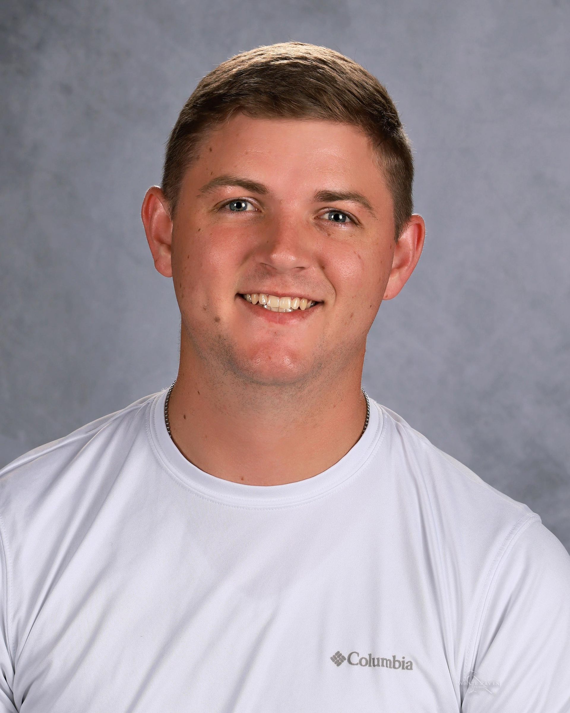 Young man in a white shirt smiling, wearing a Columbia logo. Gray background.