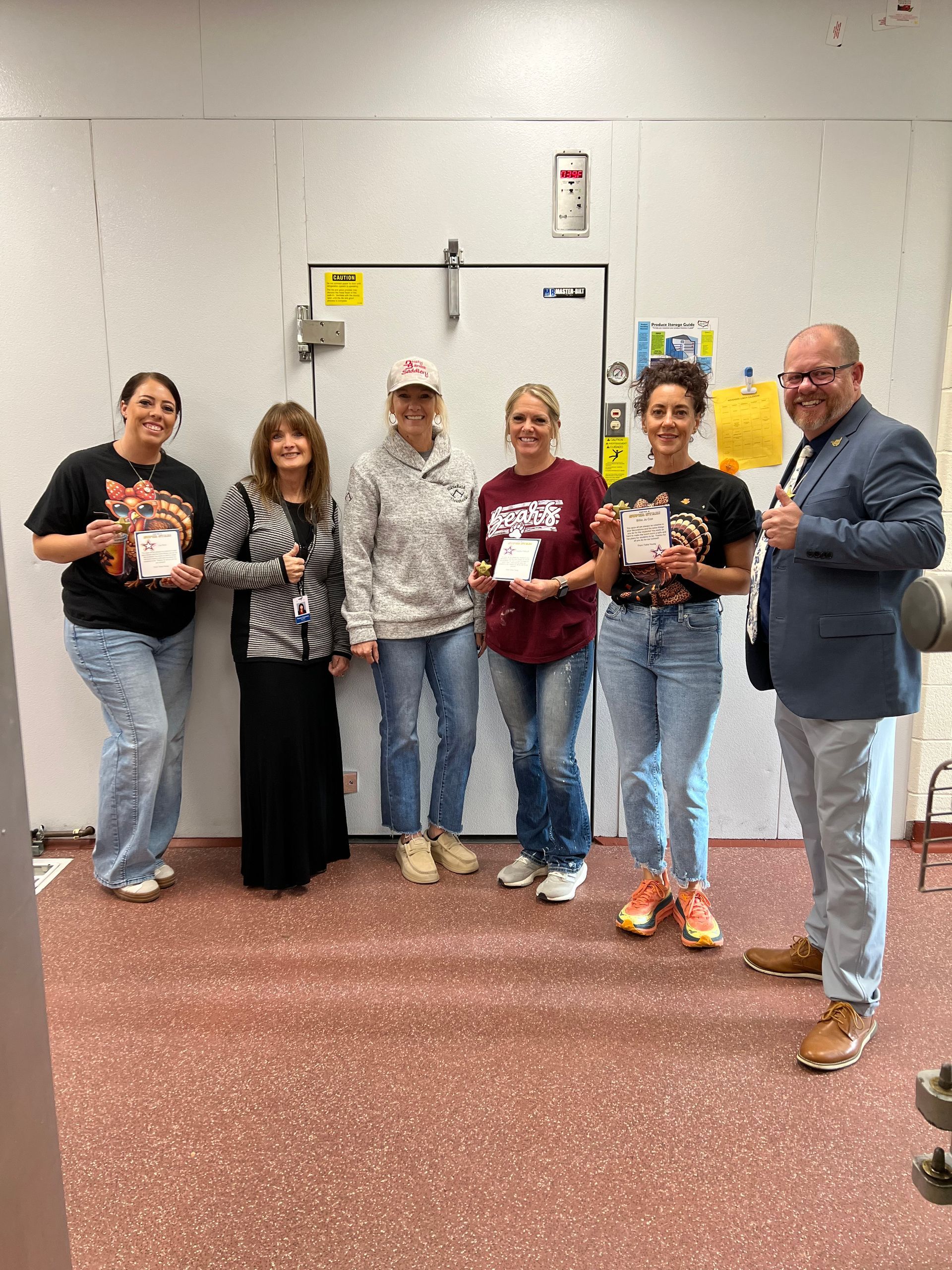 Six people stand by a closed door holding certificates. A man in a suit gives a thumbs-up. Room has red floor.