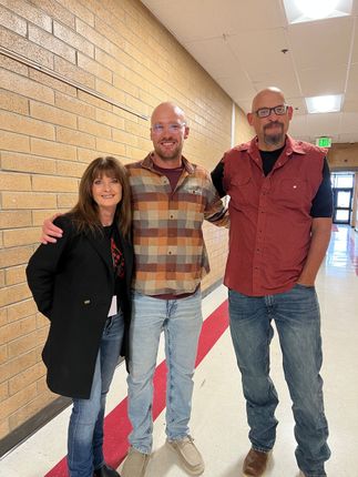 Three people stand in a hallway. A woman in a black coat and two men, one in a plaid shirt and the other in a red vest, with arms around each other.