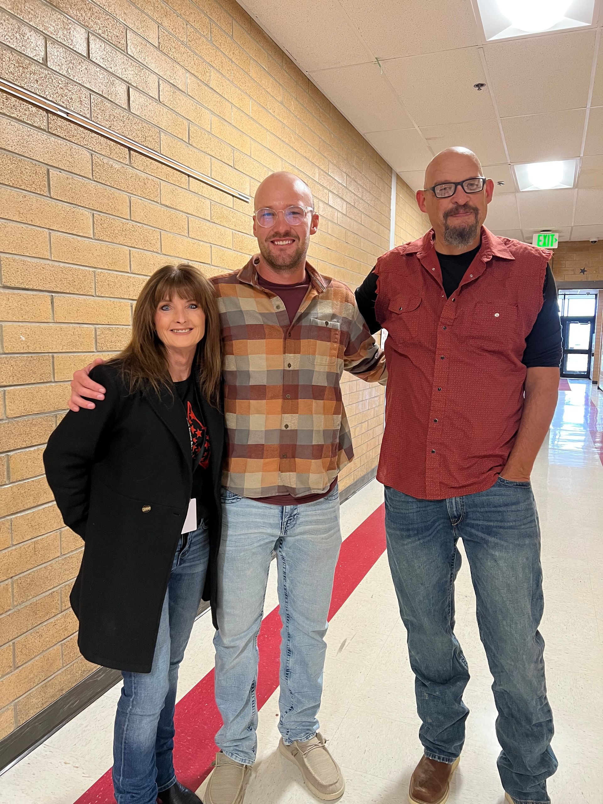 Three people pose in a hallway. A woman in a black coat and two men in button-down shirts and jeans smile, arms around each other.