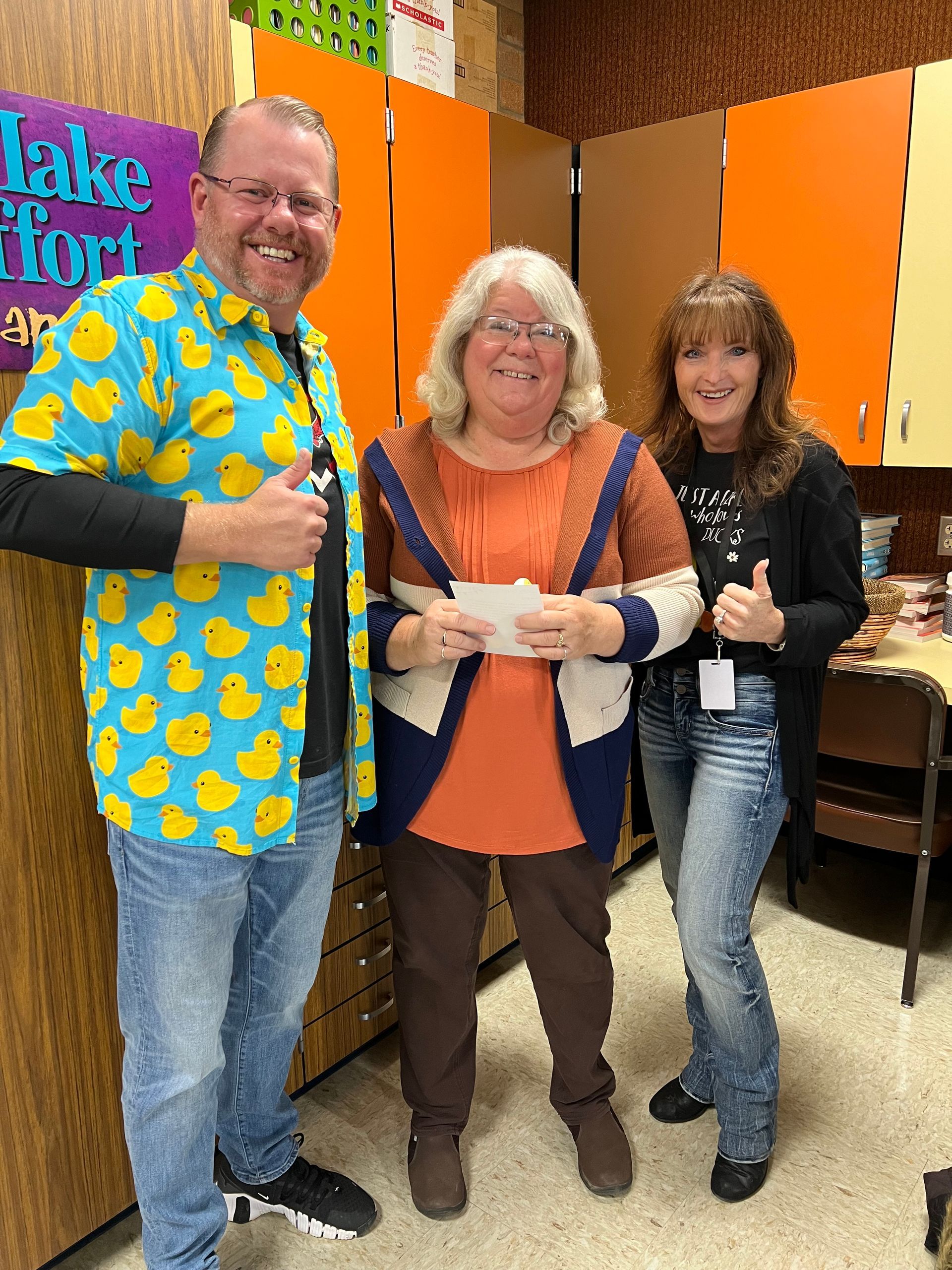 A man in a duck shirt is standing next to two women holding a check.