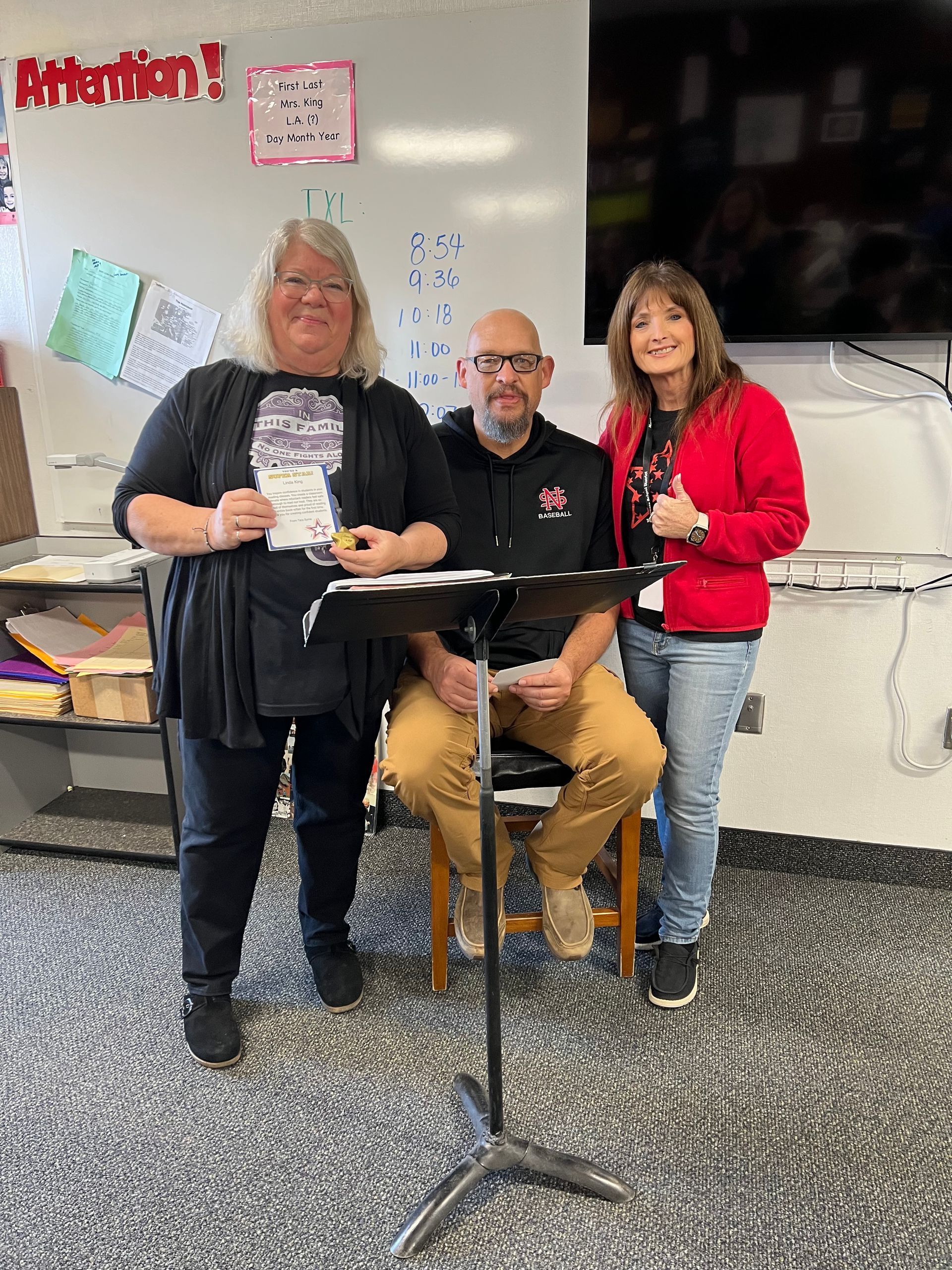 Three people in a classroom. Woman in black holds paper, man sits with papers, woman in red stands smiling.