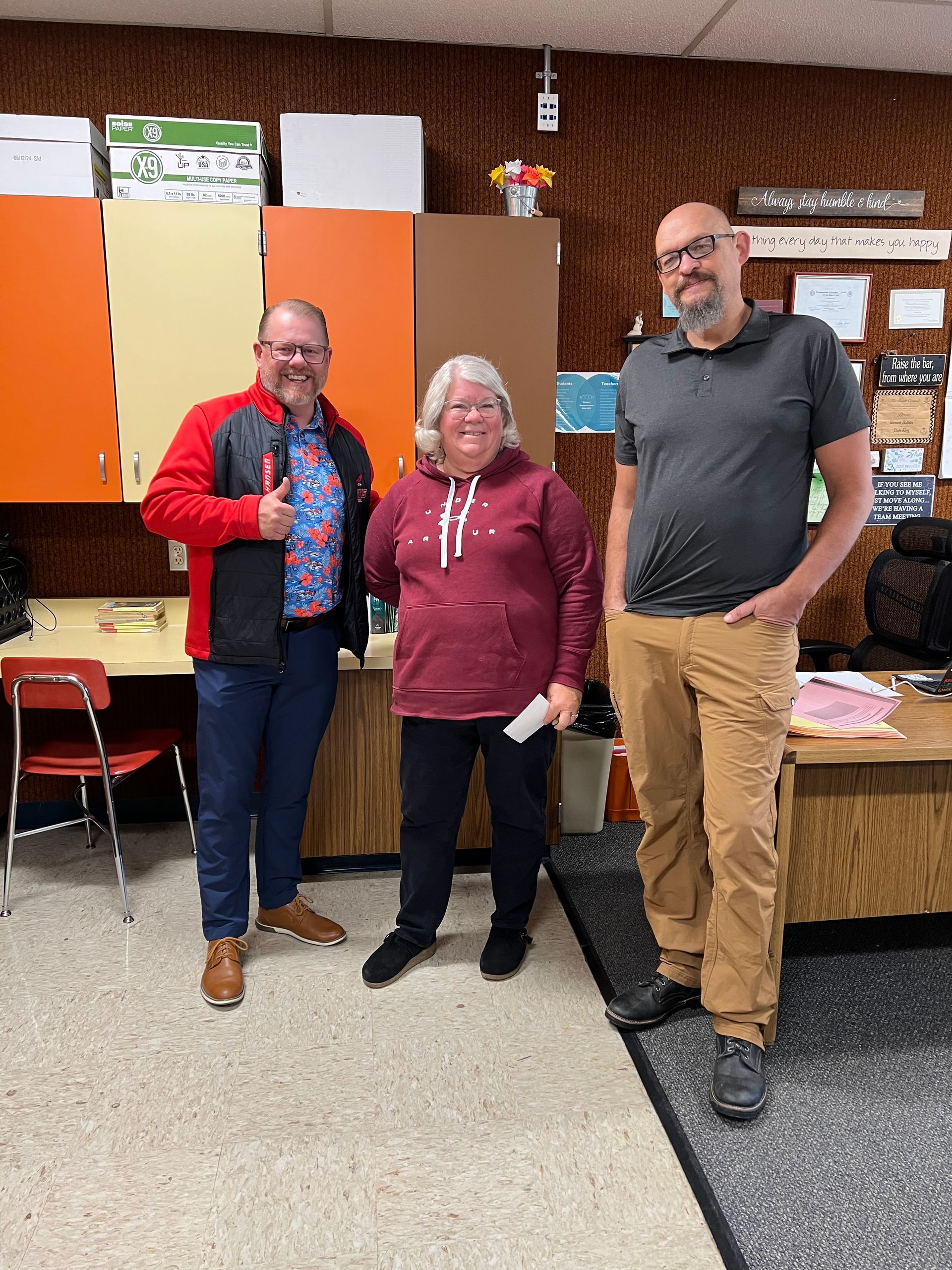 Three people standing together in a classroom, smiling in front of colorful cabinets and a desk.
