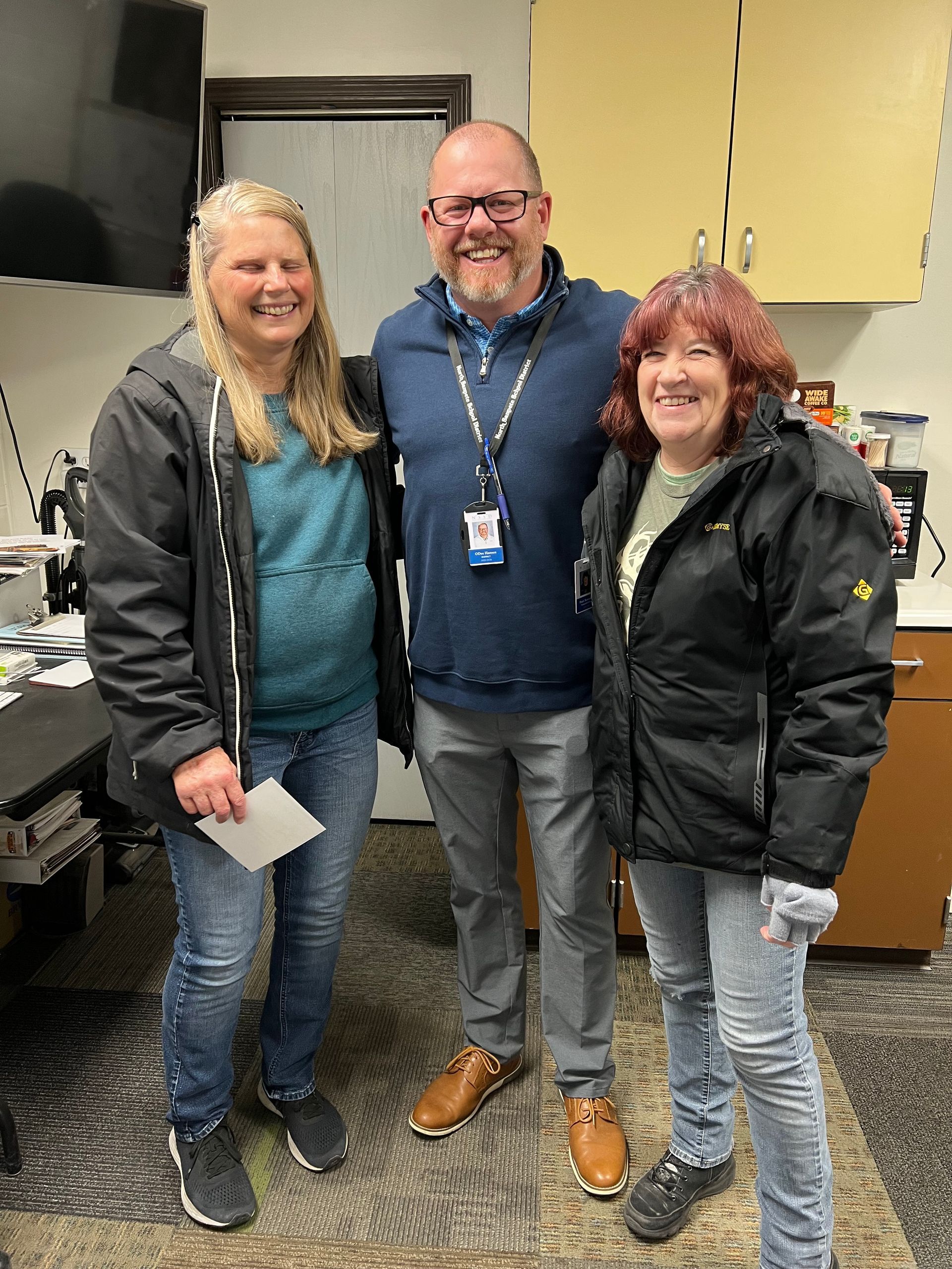 Three people smiling, standing indoors. Middle person has arm around the others. All are casually dressed.