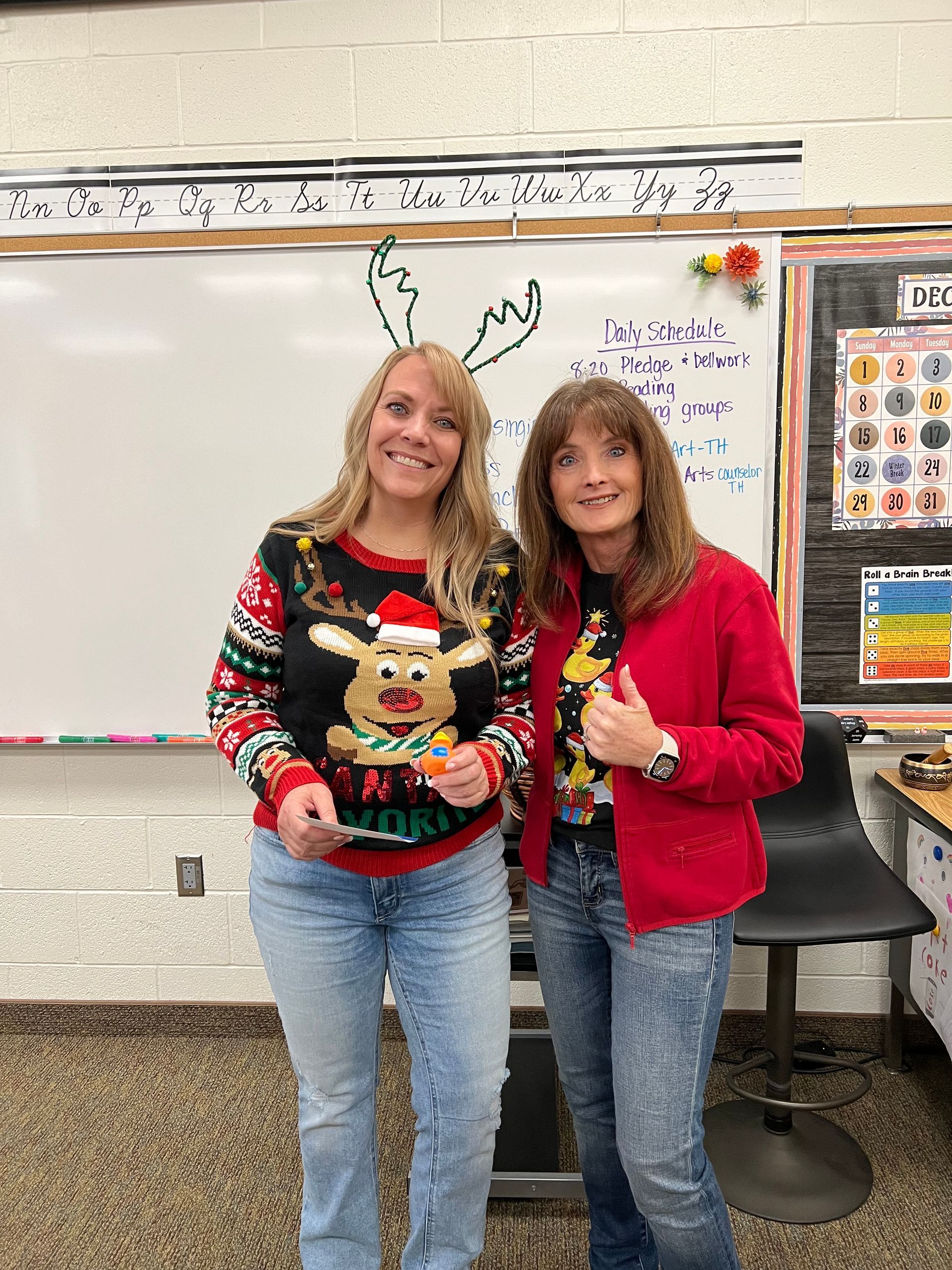 Two women are standing next to each other in a classroom wearing christmas sweaters.