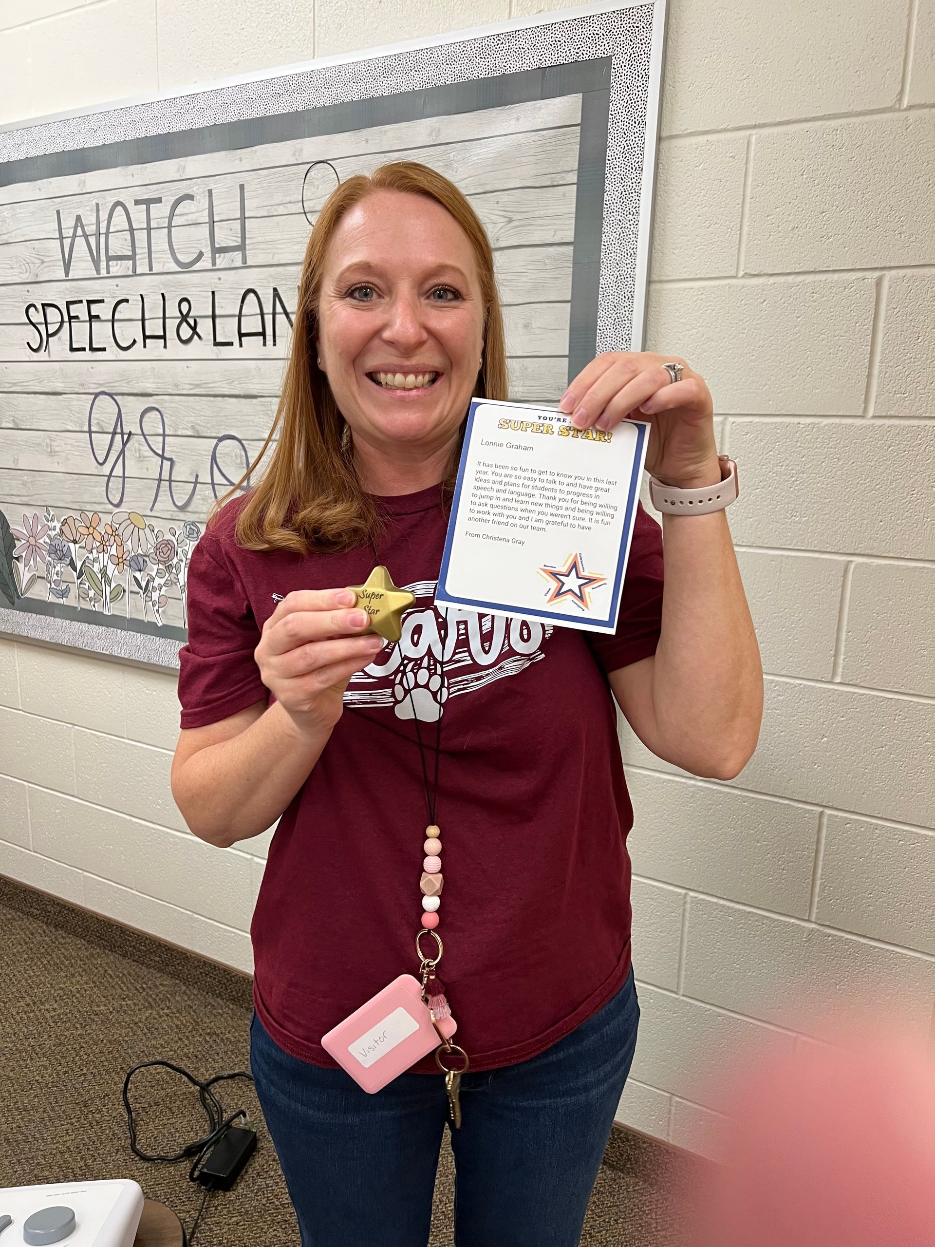 Woman with red hair smiling, holding an award and certificate in a school hallway.