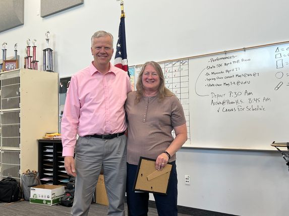 Two people stand in a classroom beside a whiteboard, smiling and holding a certificate.