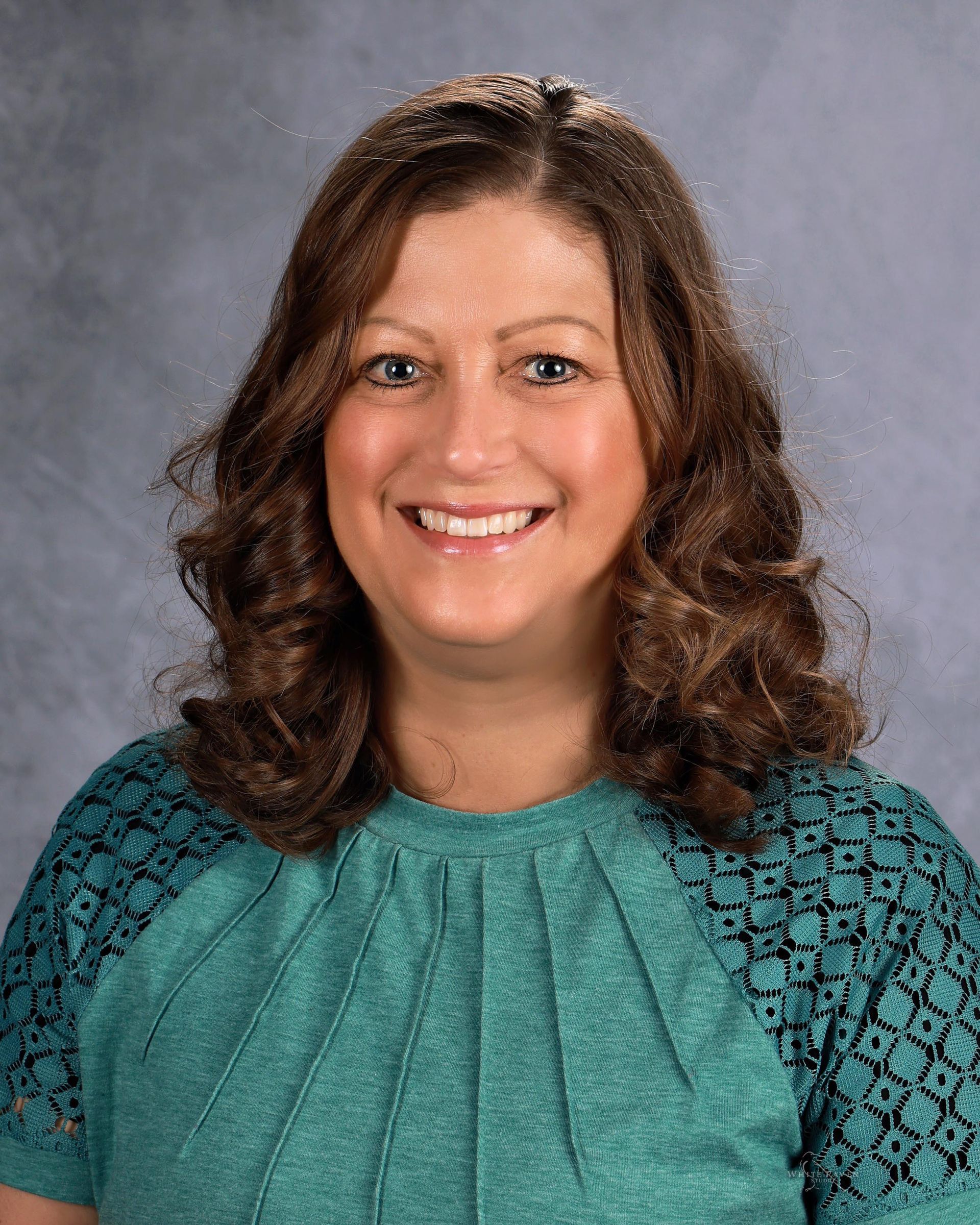 Woman with curly brown hair smiles, wearing a turquoise top, against a gray background.