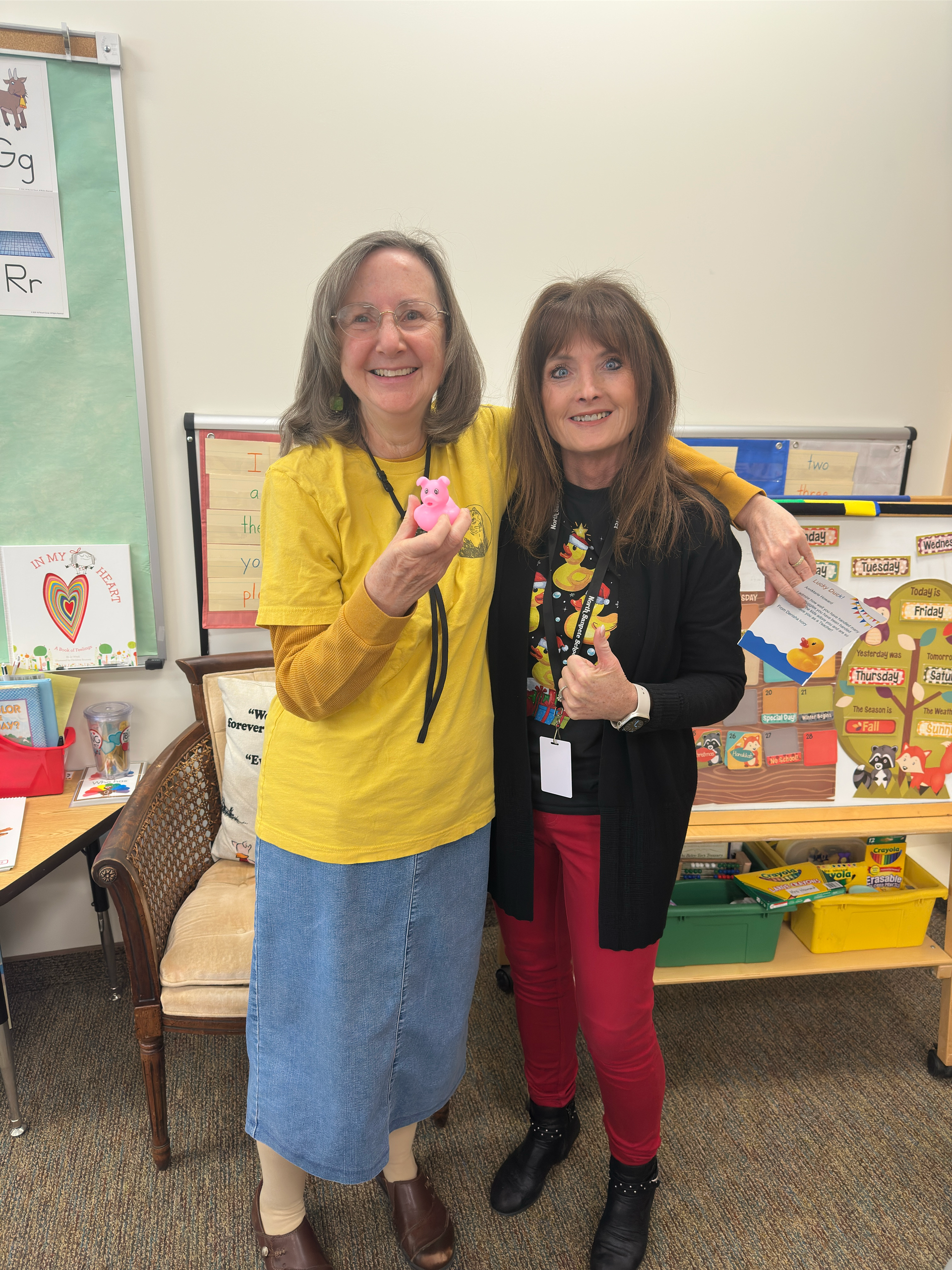 Two women are posing for a picture in a classroom . one of the women is wearing a yellow shirt.