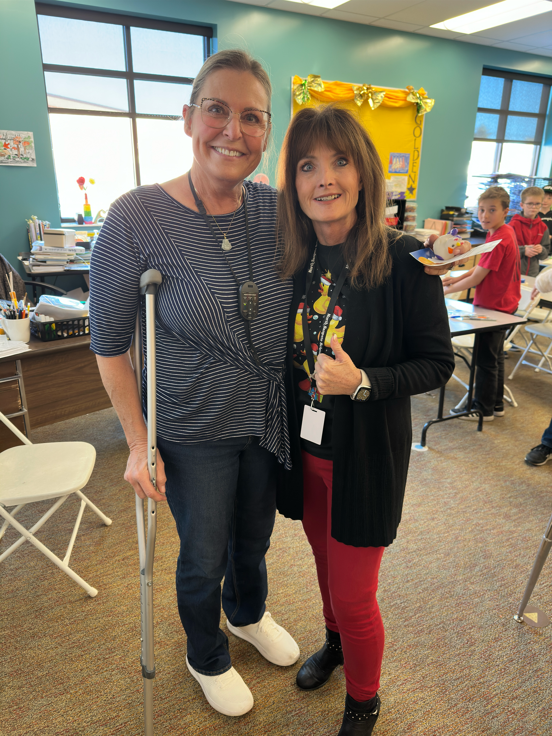 A woman with crutches is standing next to another woman in a classroom.