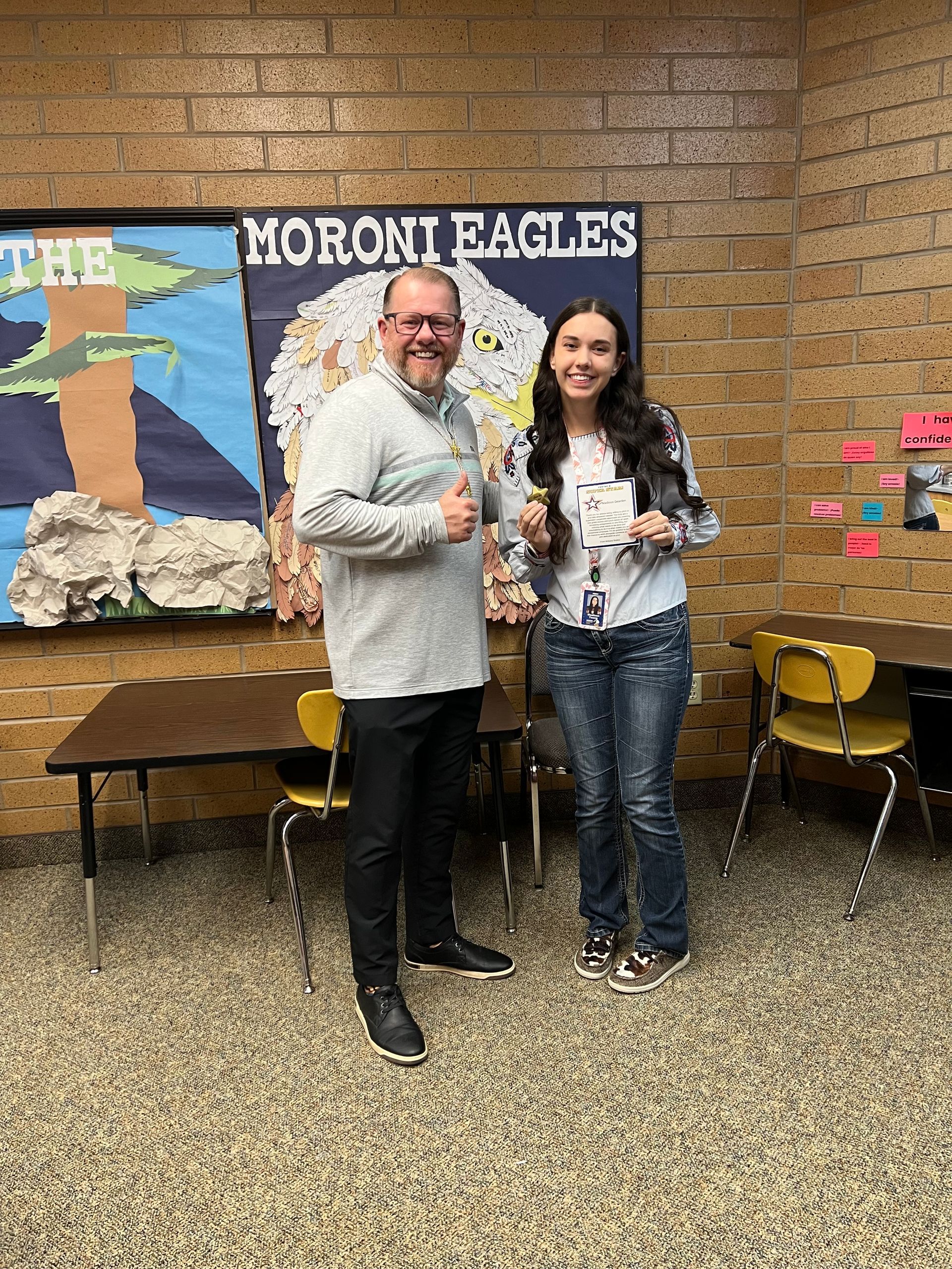 Man and woman standing in front of a school banner. They are both smiling.