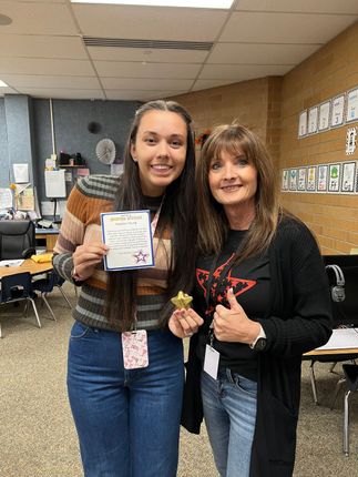 Two women smiling in a classroom, one holding a note and a small item, the other giving a thumbs-up.