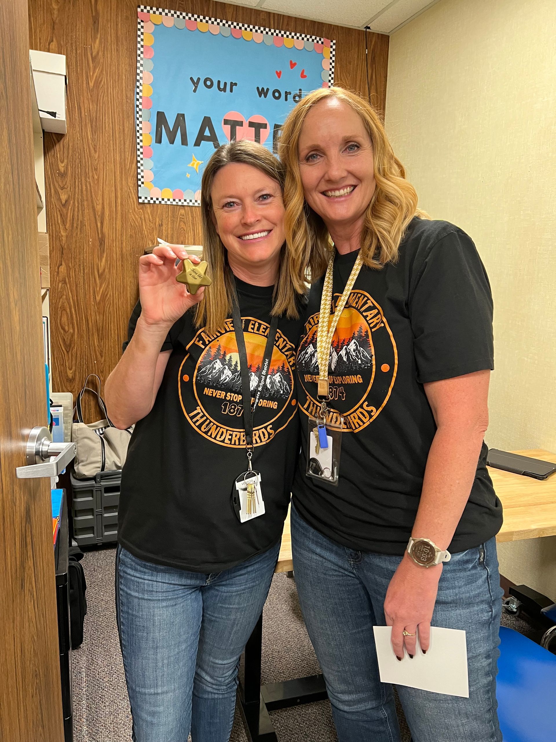 Two women in matching black shirts and jeans smile, posing in a classroom. One holds a gold item.