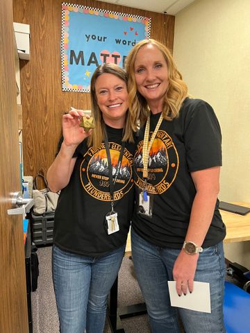 Two women in matching shirts smile, one holds a small award. Brown-paneled wall with a 