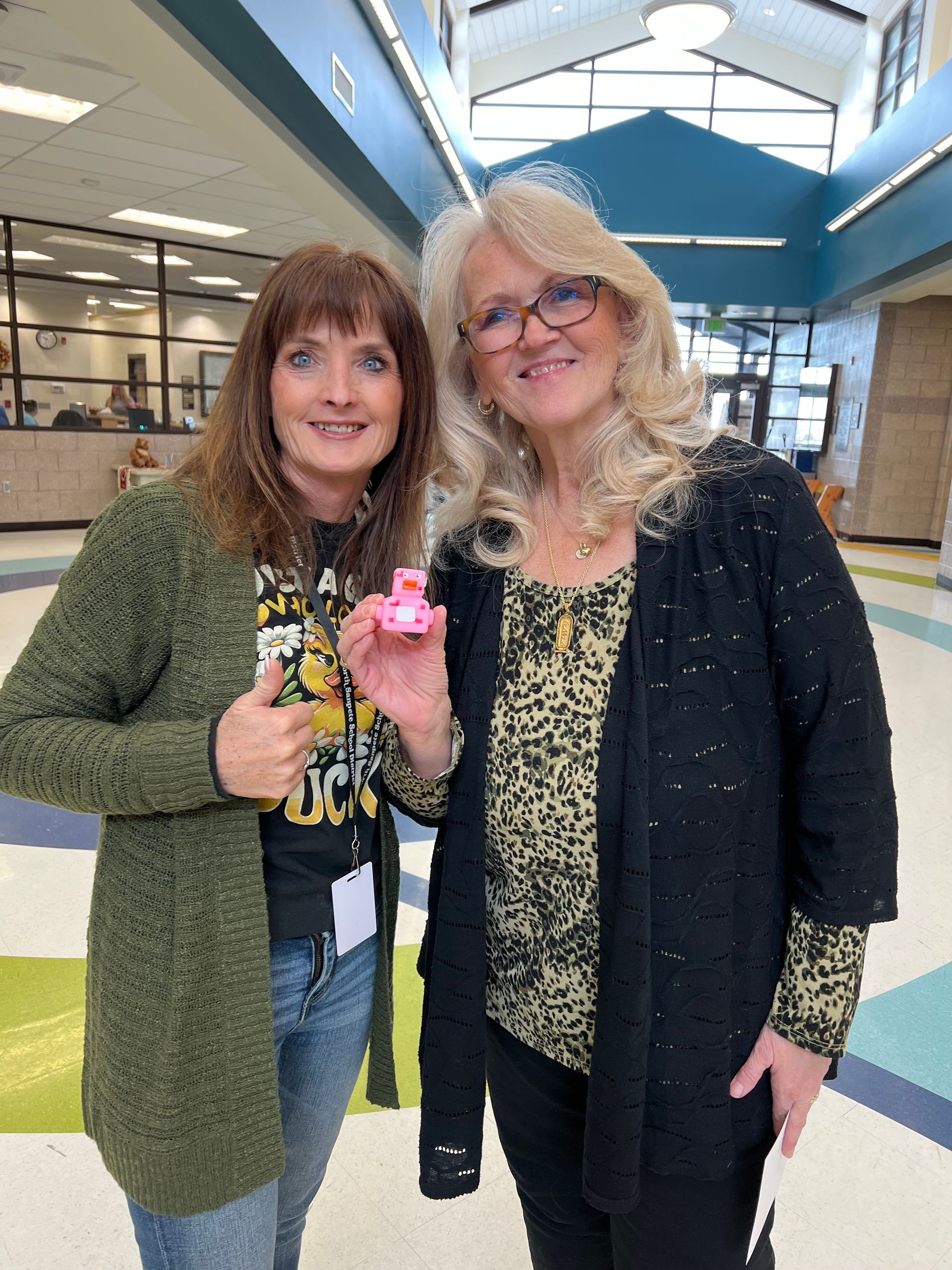 Two women are standing next to each other in a hallway holding a pink heart.