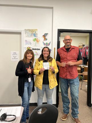 Three adults stand by a door: woman in a yellow jacket holds a card, woman gives thumbs-up, man in red shirt.