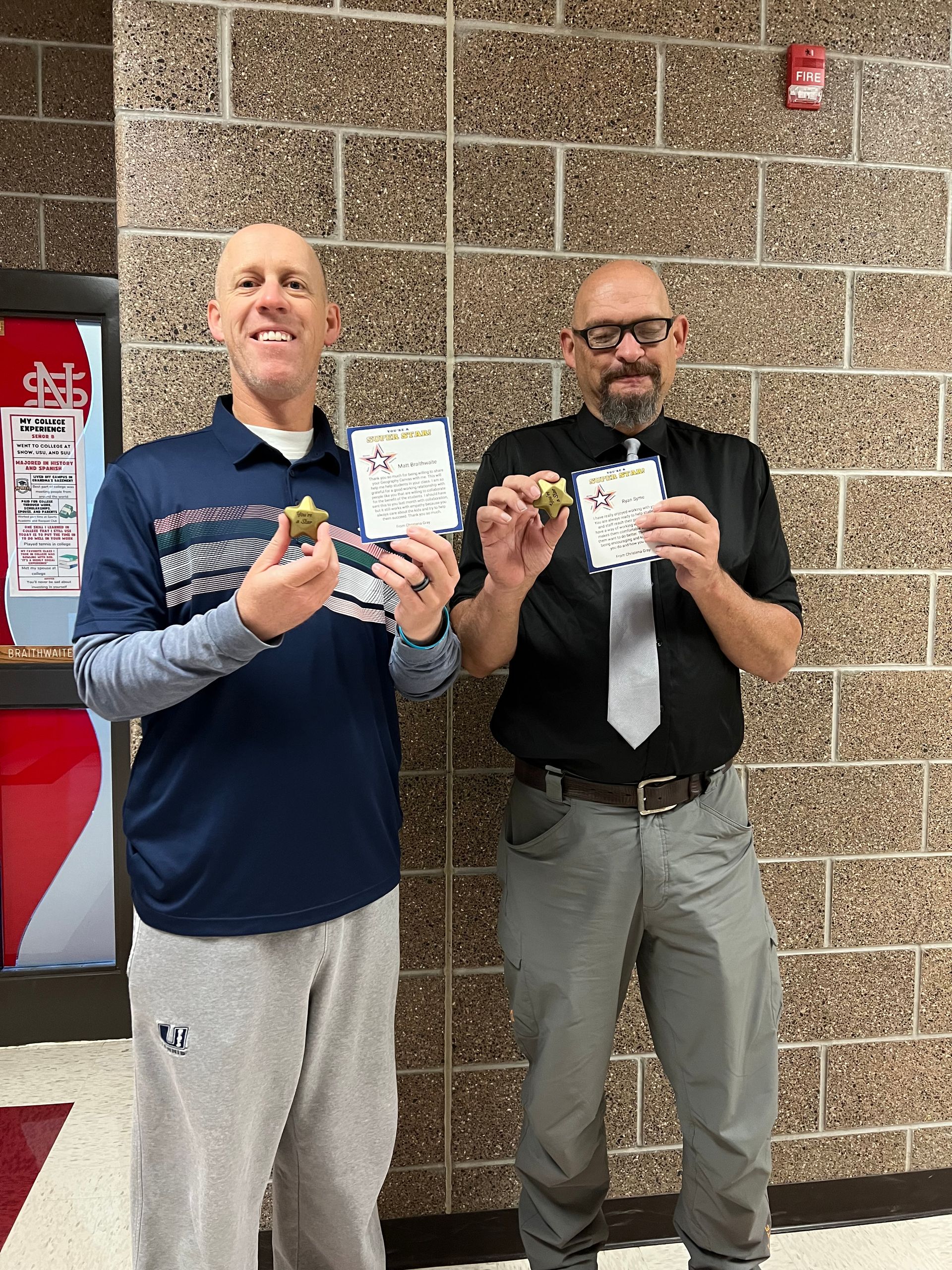Two men in front of brick wall hold small items and cards, smiling. One in athletic wear, other in shirt and tie.