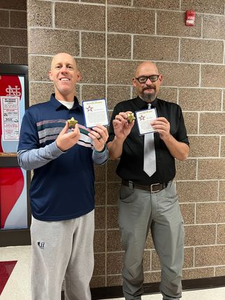 Two men holding small trophies and papers, smiling in a school hallway with a soda machine visible.
