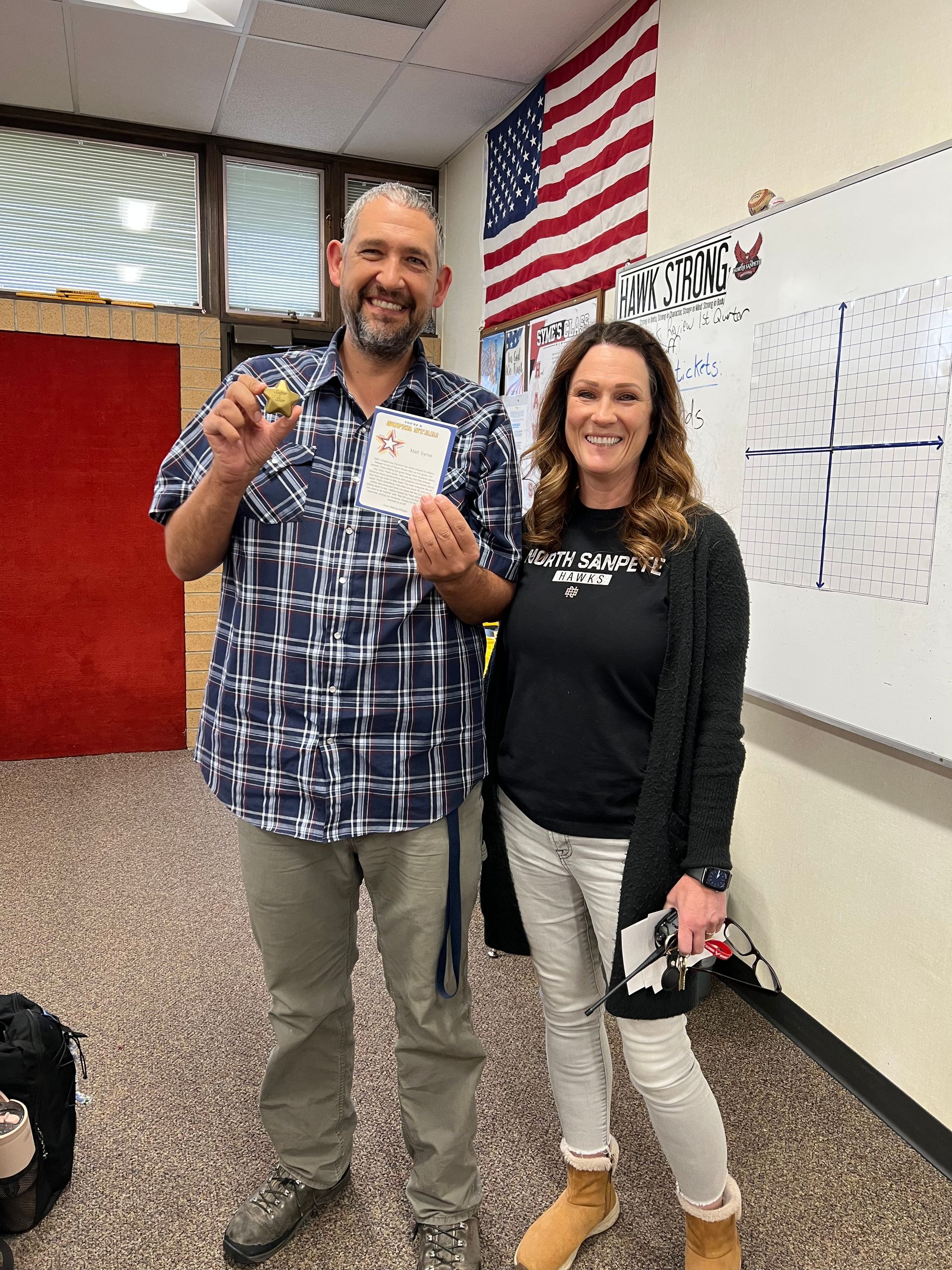 Two people in a classroom: man holds a coin and card, woman smiles. American flag and whiteboard in background.